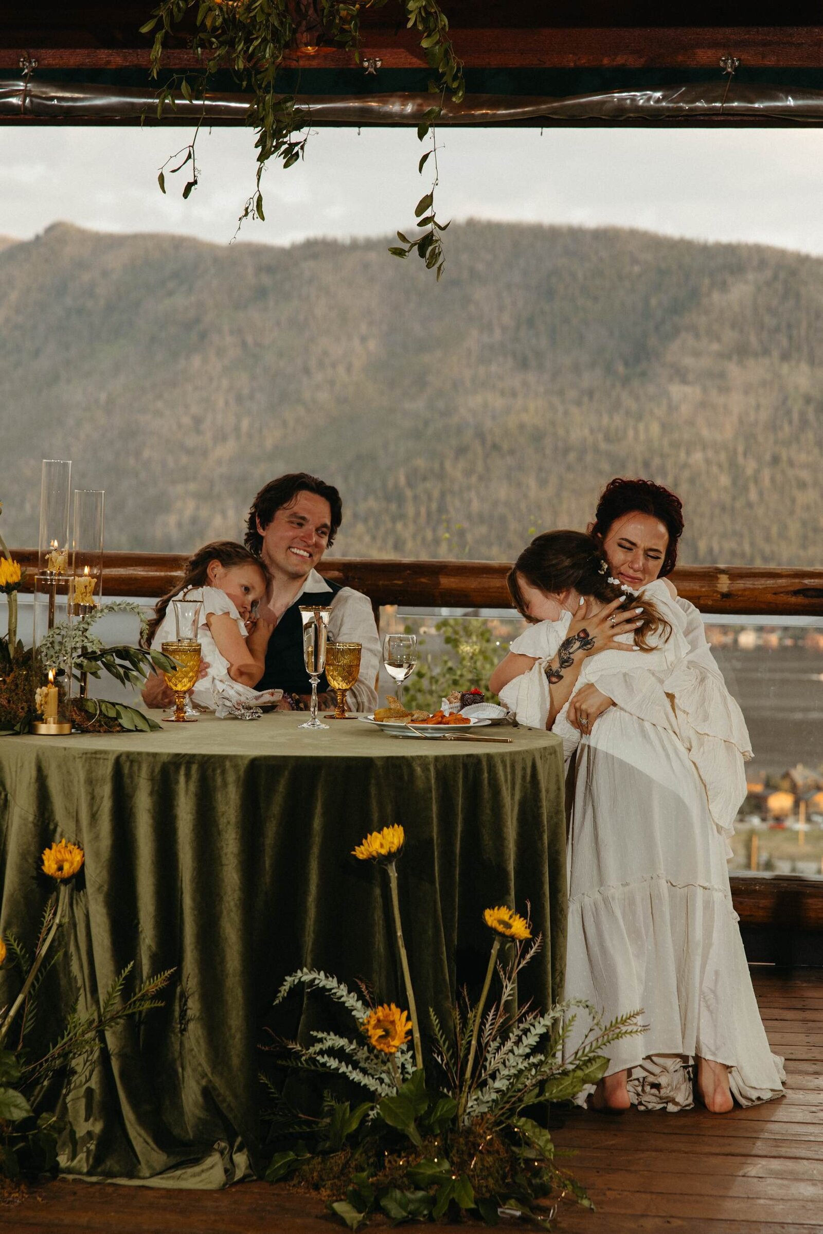 A wedding couple sits at their green velvet sweetheart table flanked by floral arrangements featuring sunflowers, hugging their daughters who are sitting on their laps