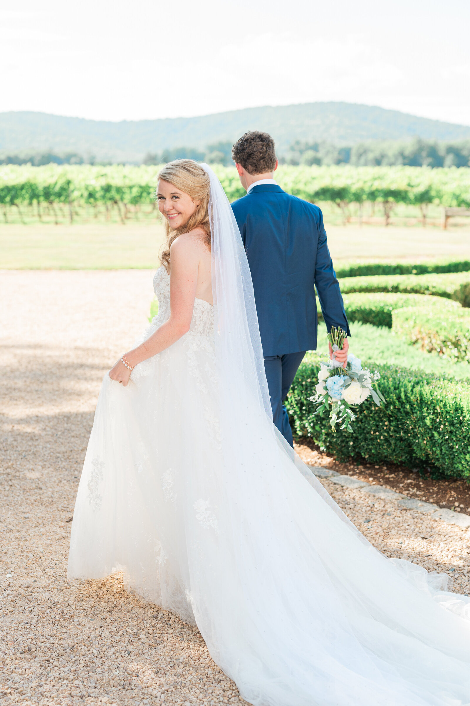 A wedding couple walking along a gravel path as one looks back over their shoulder smiling