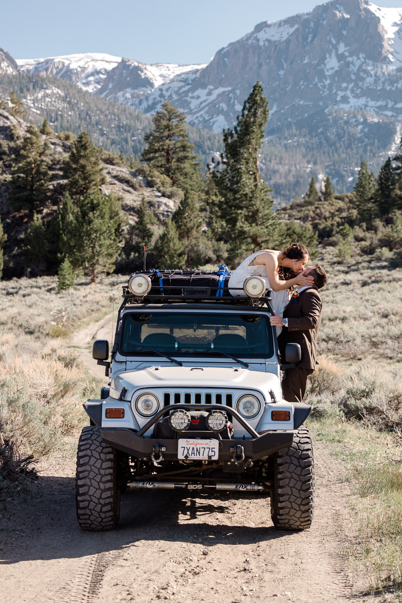 A bride sits on top a jeep outfitted for off roading, and leans down to kiss her groom while on forest service road.