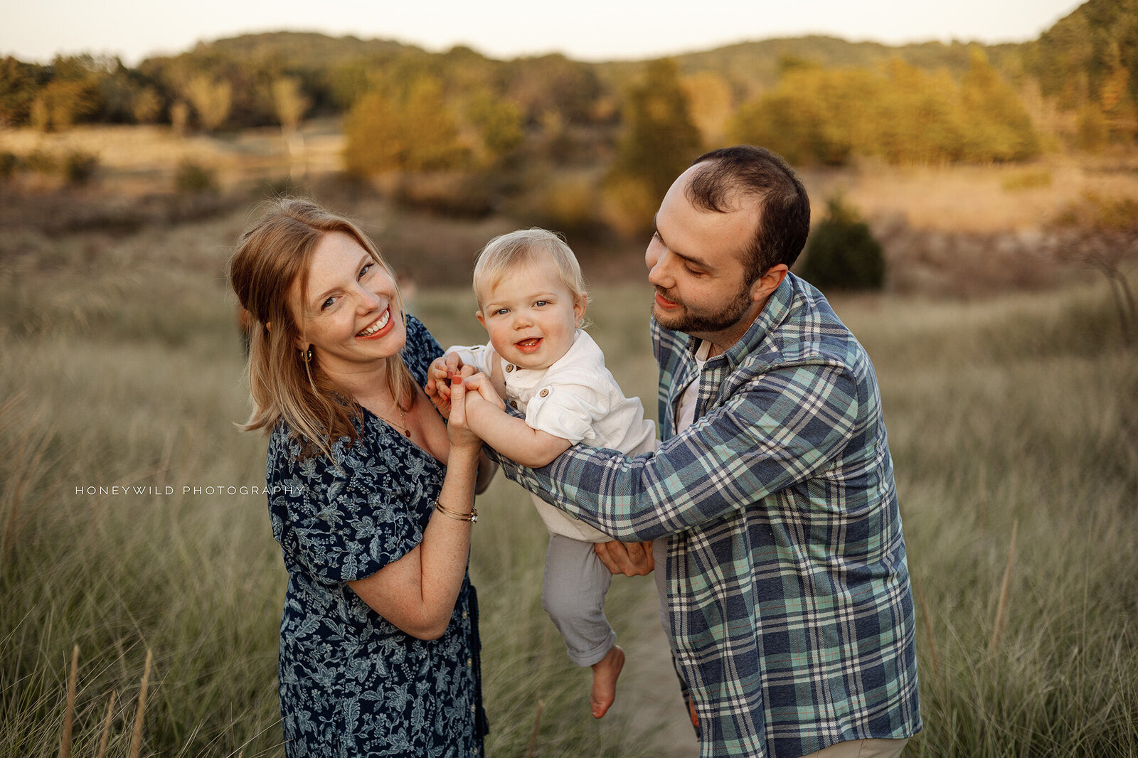 A happy family of three enjoying an outdoor photography session in Grand Rapids, Michigan, with a mother and father holding their smiling baby in a scenic natural setting.