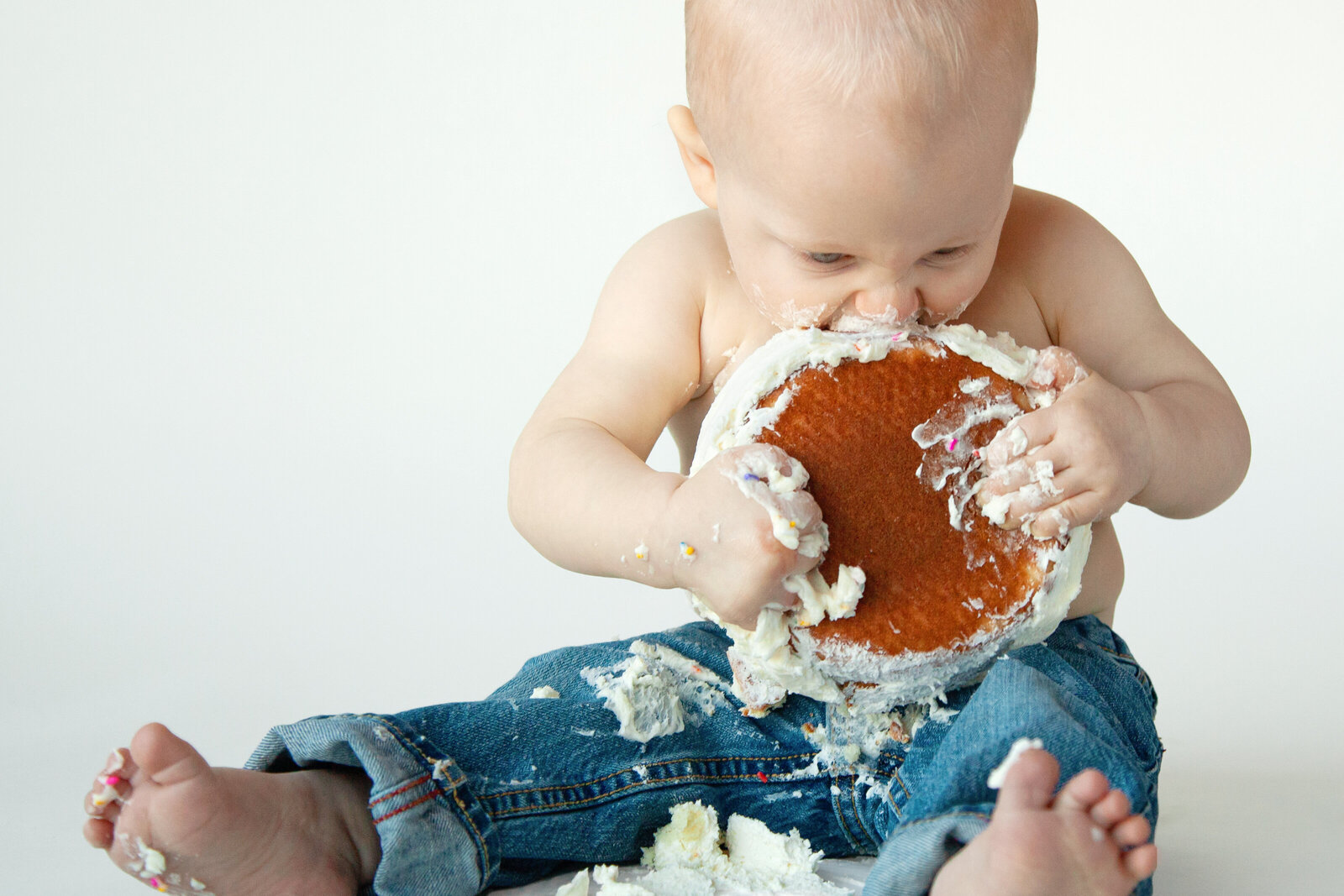 One year old boy in jeans picking up smash cake to eat it.