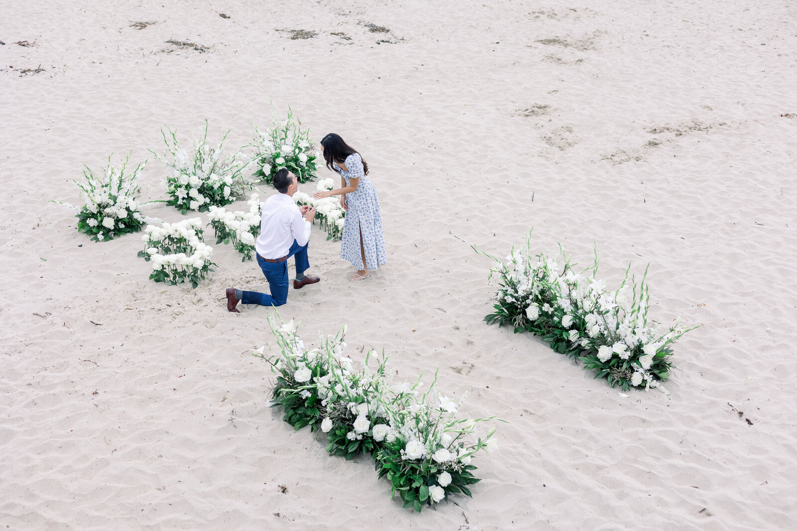 surprise proposal pictures at half moon bay beach with white florals set up as the man in on one knee asking his girlfriend to marry him photographed by wedding photographer bay area