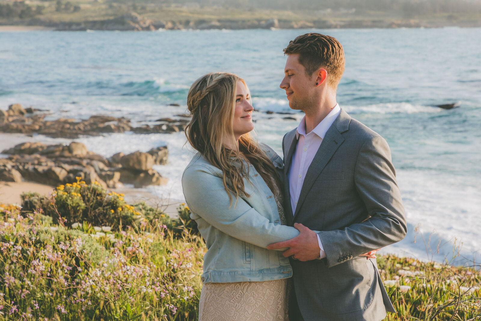 A bride wearing a denium jacket looks at her groom with the ocean behind them in Carmel.