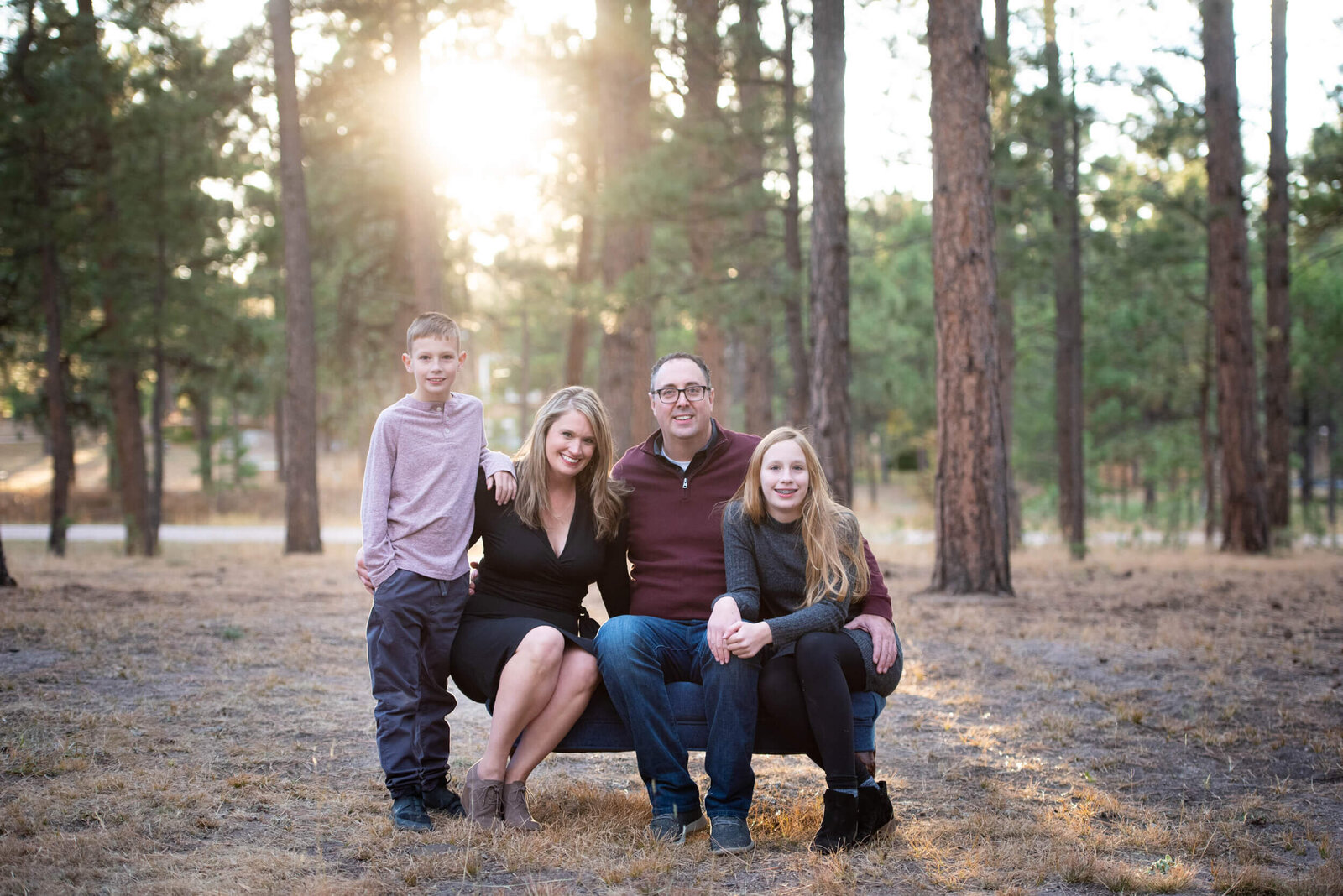 A family of 4 sit on a park bench in a pine forest at sunset