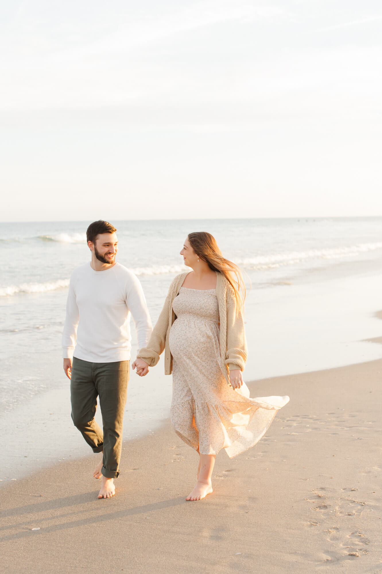 Pregnant couple walks hand in hand along the beach near the shoreline during golden hour