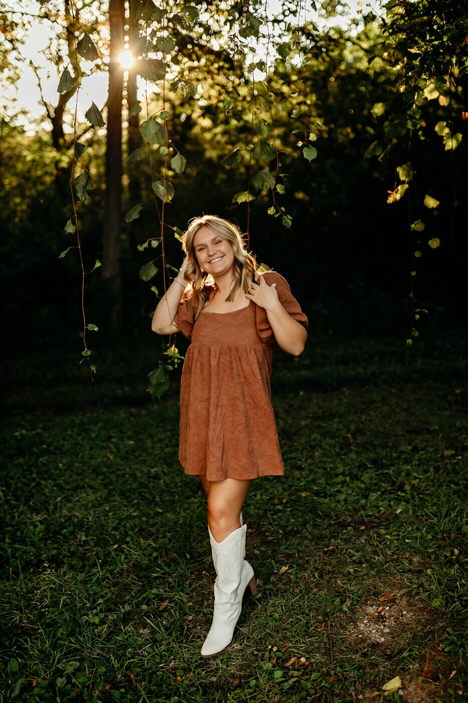 A person in a brown dress and white boots stands in a sunlit forest setting, smiling while holding hanging vines. The sunlight filters through the trees, illuminating the scene.