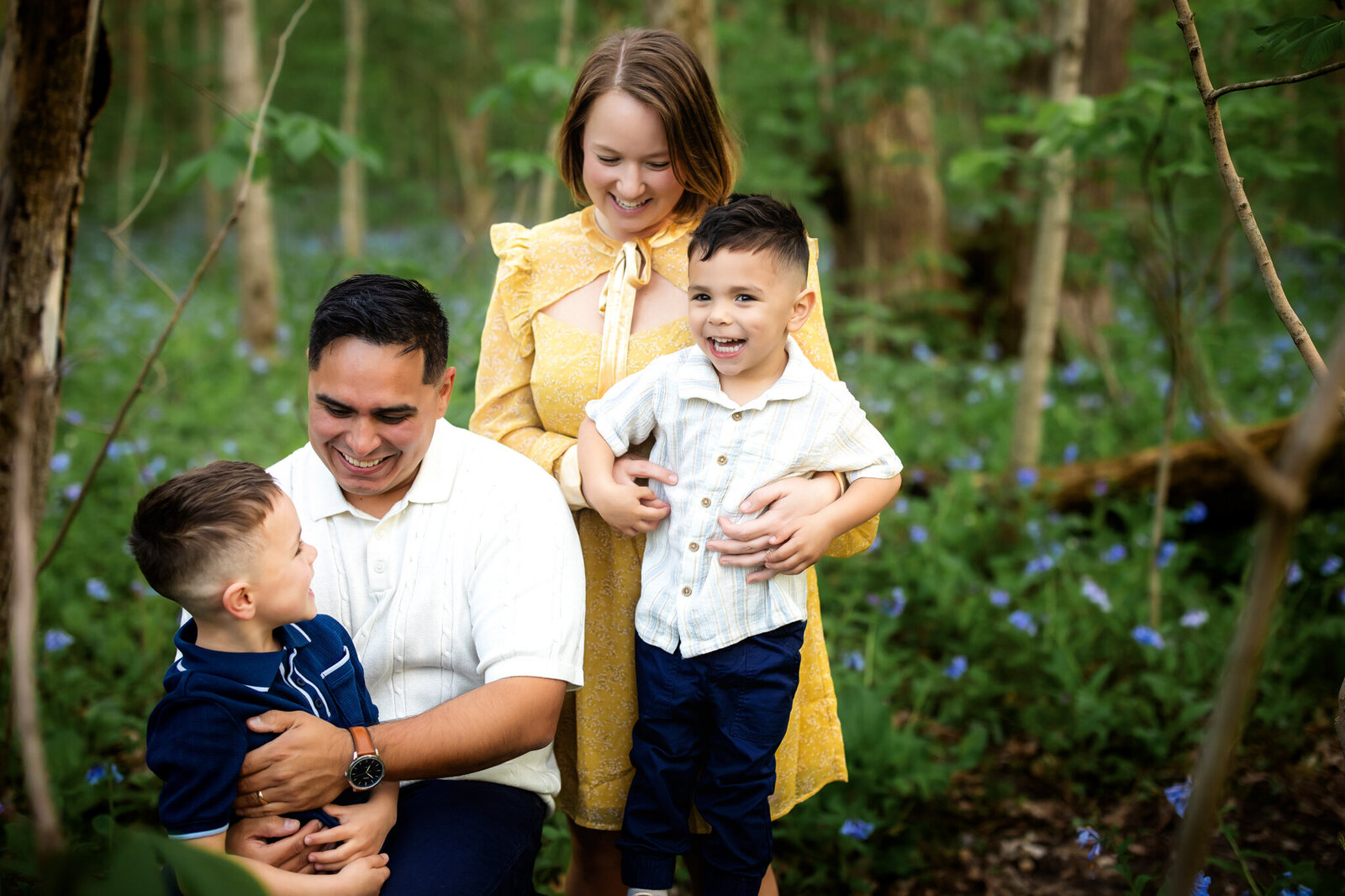 A smiling family of four poses in a lush green forest, captured by a Bloomington family photographer. The mother in a yellow dress hugs her two sons from behind, while the father kneels, smiling warmly at one of the boys.