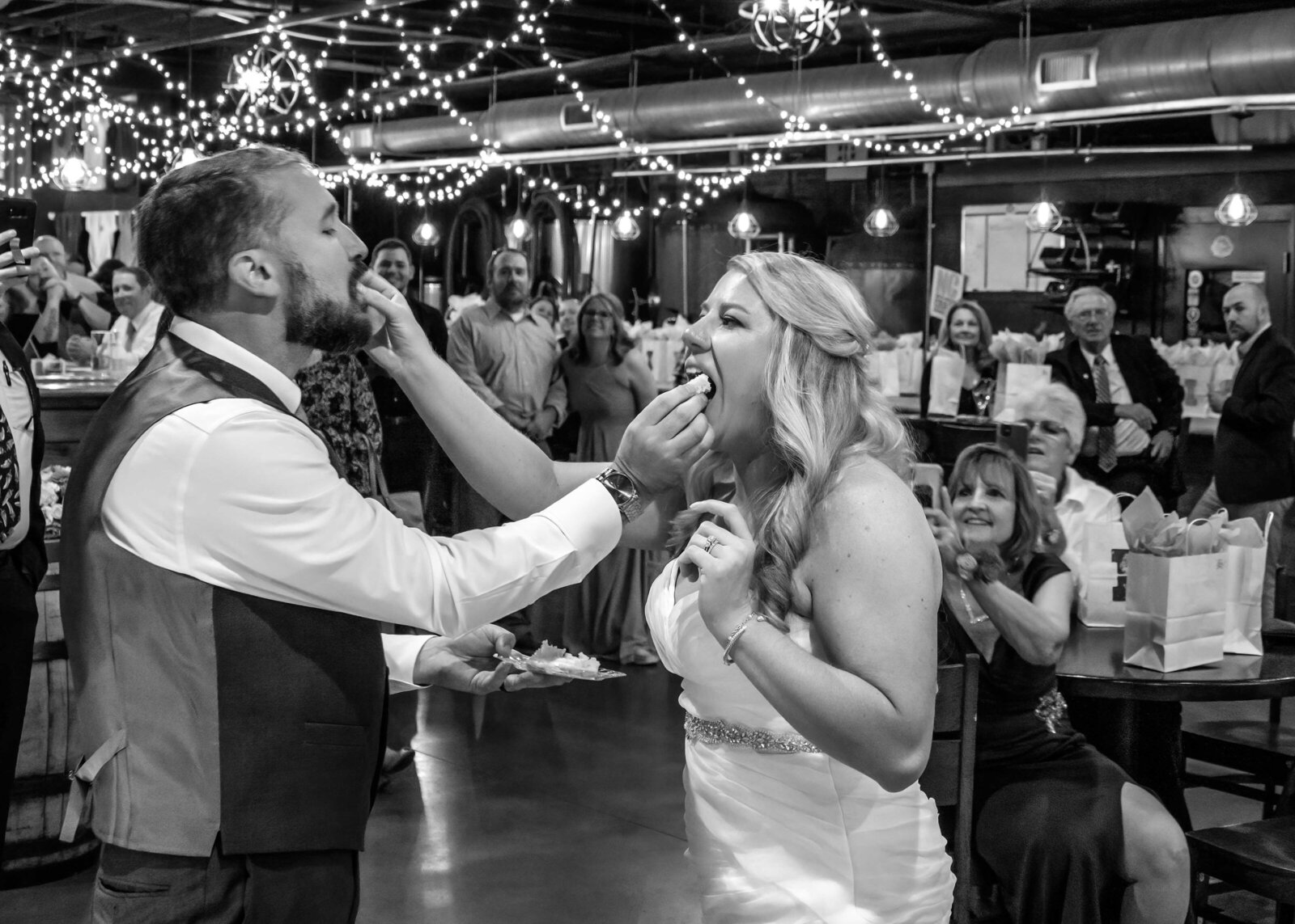 Black-and-white photo of the bride and groom sharing a joyful cake-feeding moment surrounded by guests and twinkling string lights, captured during a Wilmington, NC wedding reception.