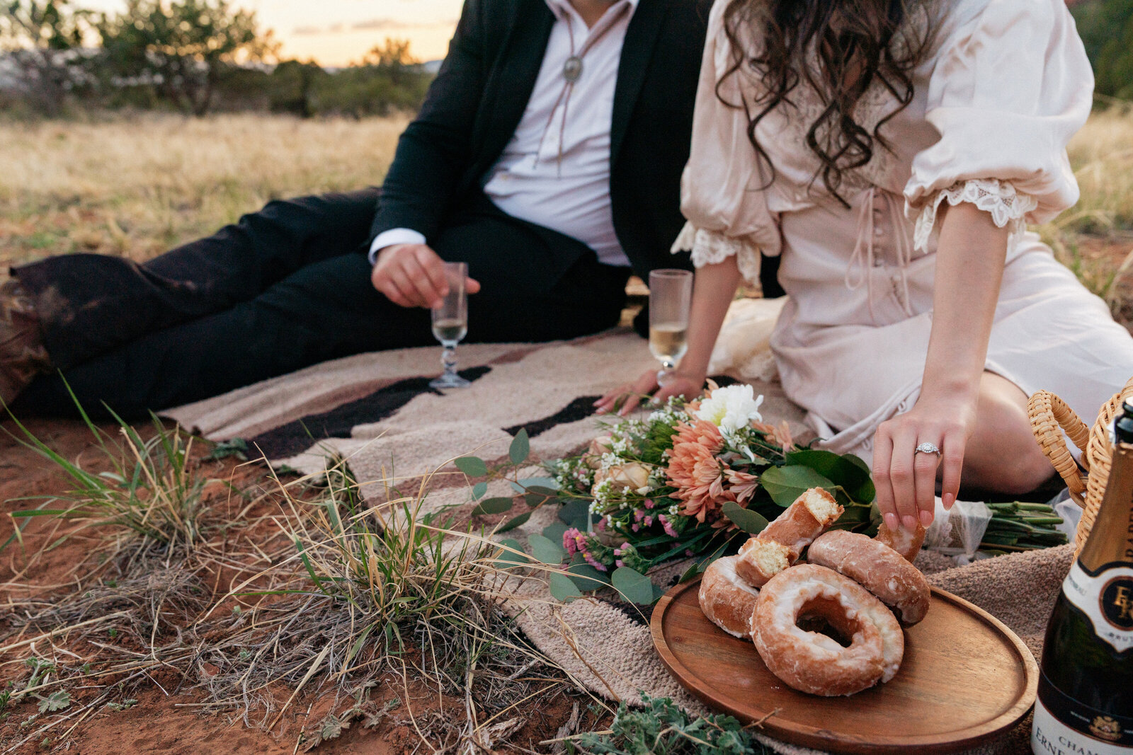 Bride and groom enjoying a romantic elopement picnic at Bear Mountain in Sedona, Arizona, featuring a cozy blanket setup with donuts, champagne, and a wedding bouquet at sunset.