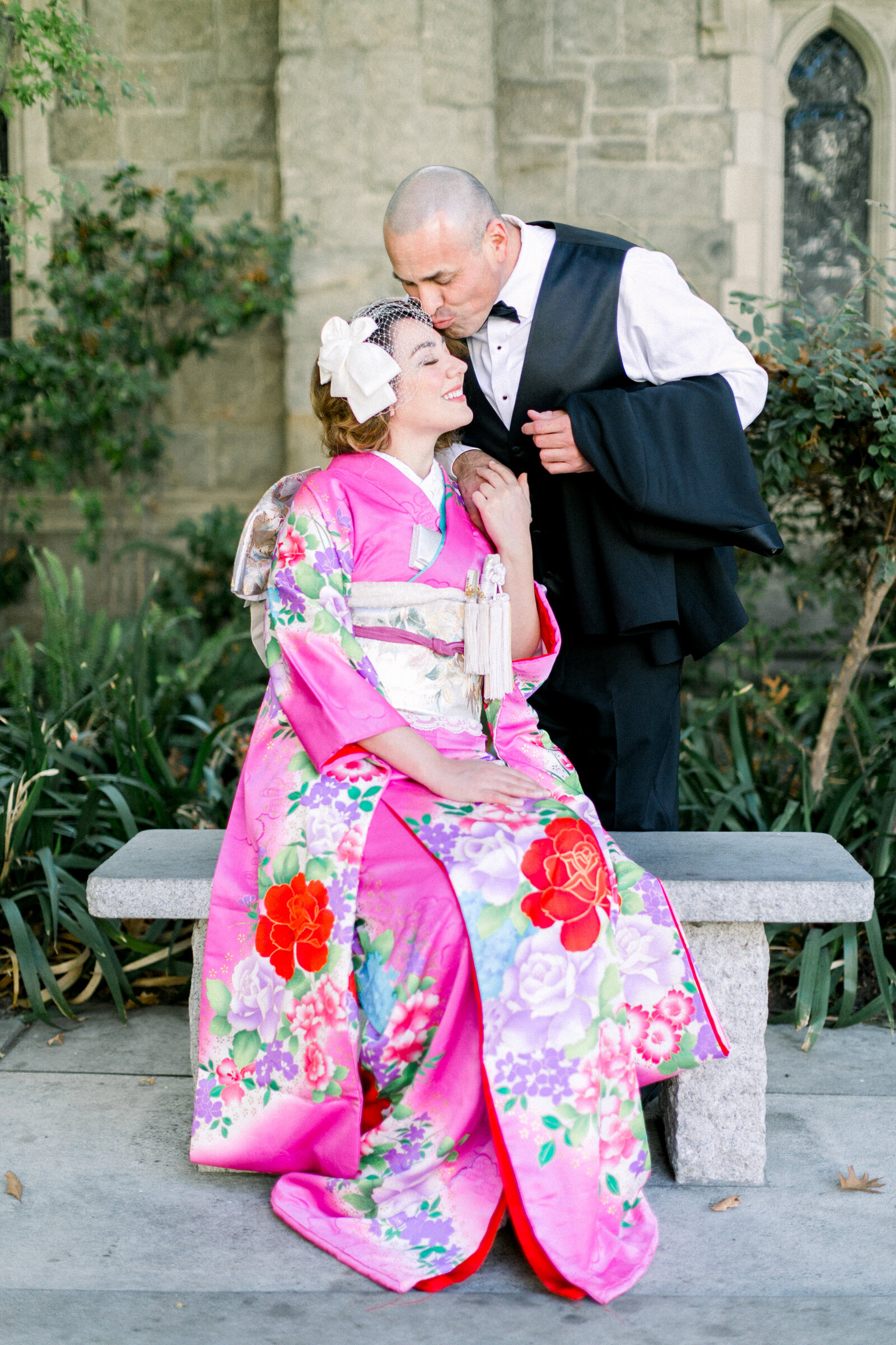Bride in vivid pink wedding kimono rental sitting on a stone bench while groom kisses her forehead at All Saints Episcopal Church, Pasadena California | オールセインツ教会の石のベンチでピンク引き振袖レンタル花嫁の額にキスする新郎