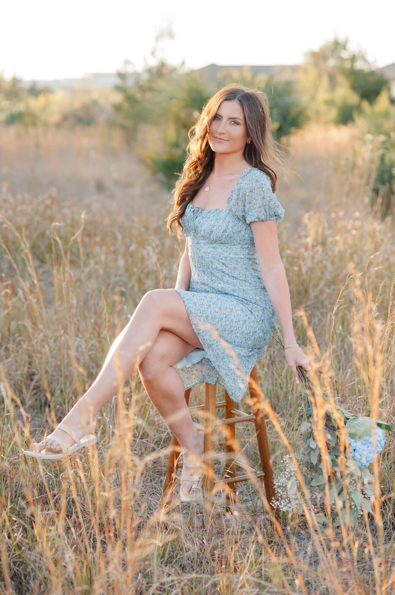 Stunning senior wearing a blue floral dress sitting in tall grass on a bar stool holding florals
