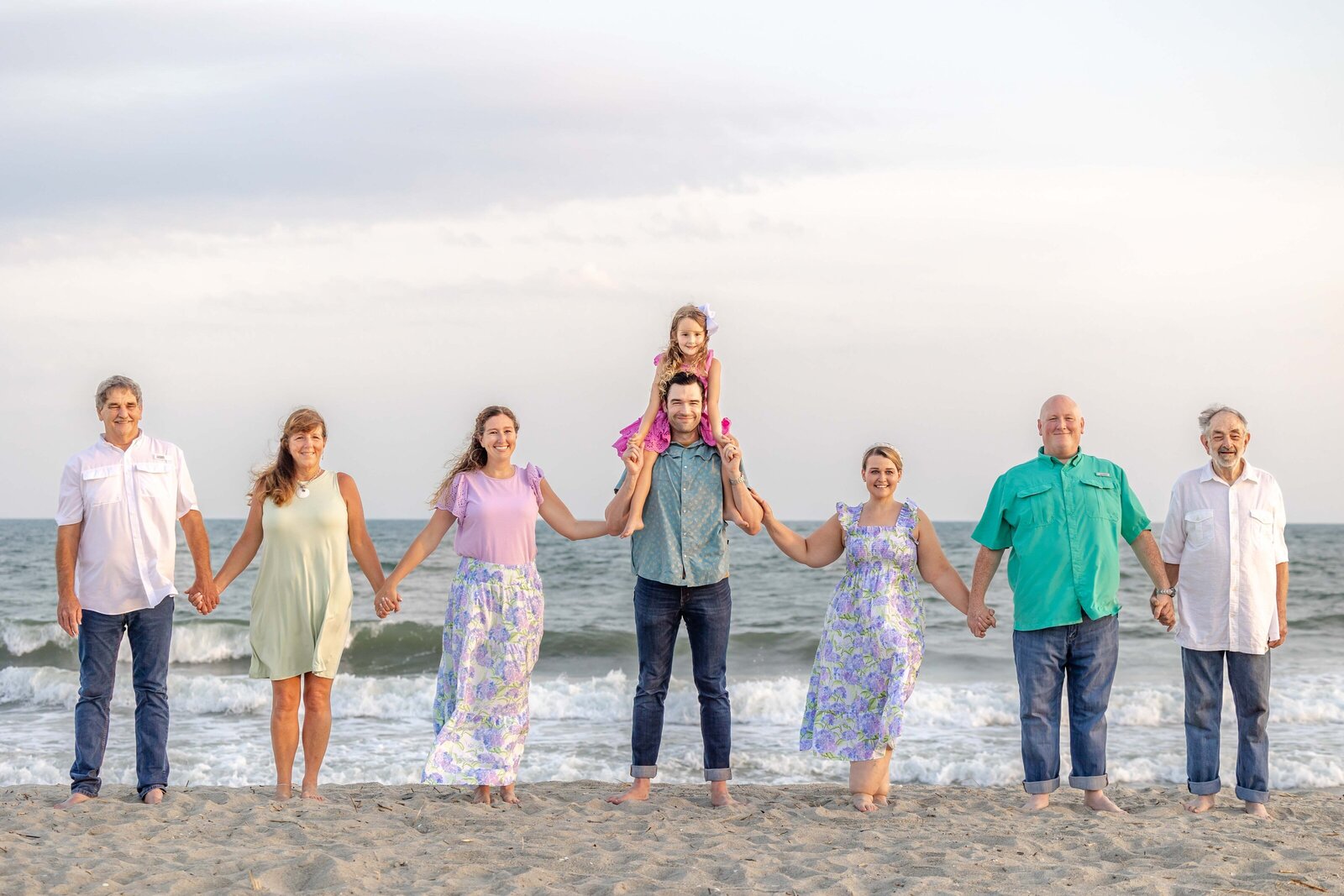 An extended family stands hand in hand on the beach, with one child on shoulders, dressed in pastel and teal outfits. The ocean waves provide a beautiful backdrop, perfect for North Carolina family photography, capturing joyful and connected moments.