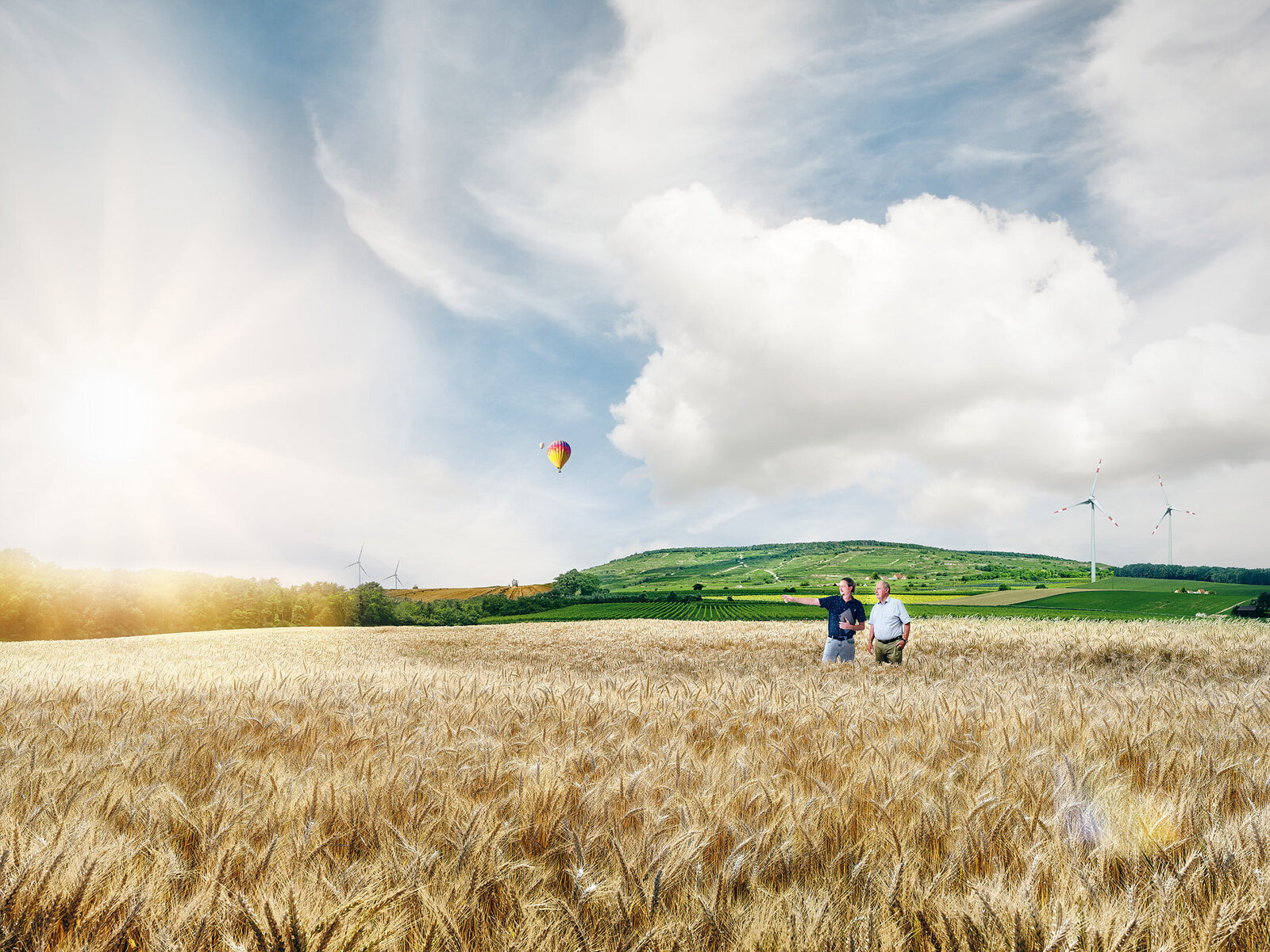 Zwei Personen bei der Landschaftsplanung in einem Weizenfeld mit Windrädern und Heißluftballon – nachhaltige Agrar- und Energiethemen im Fokus.