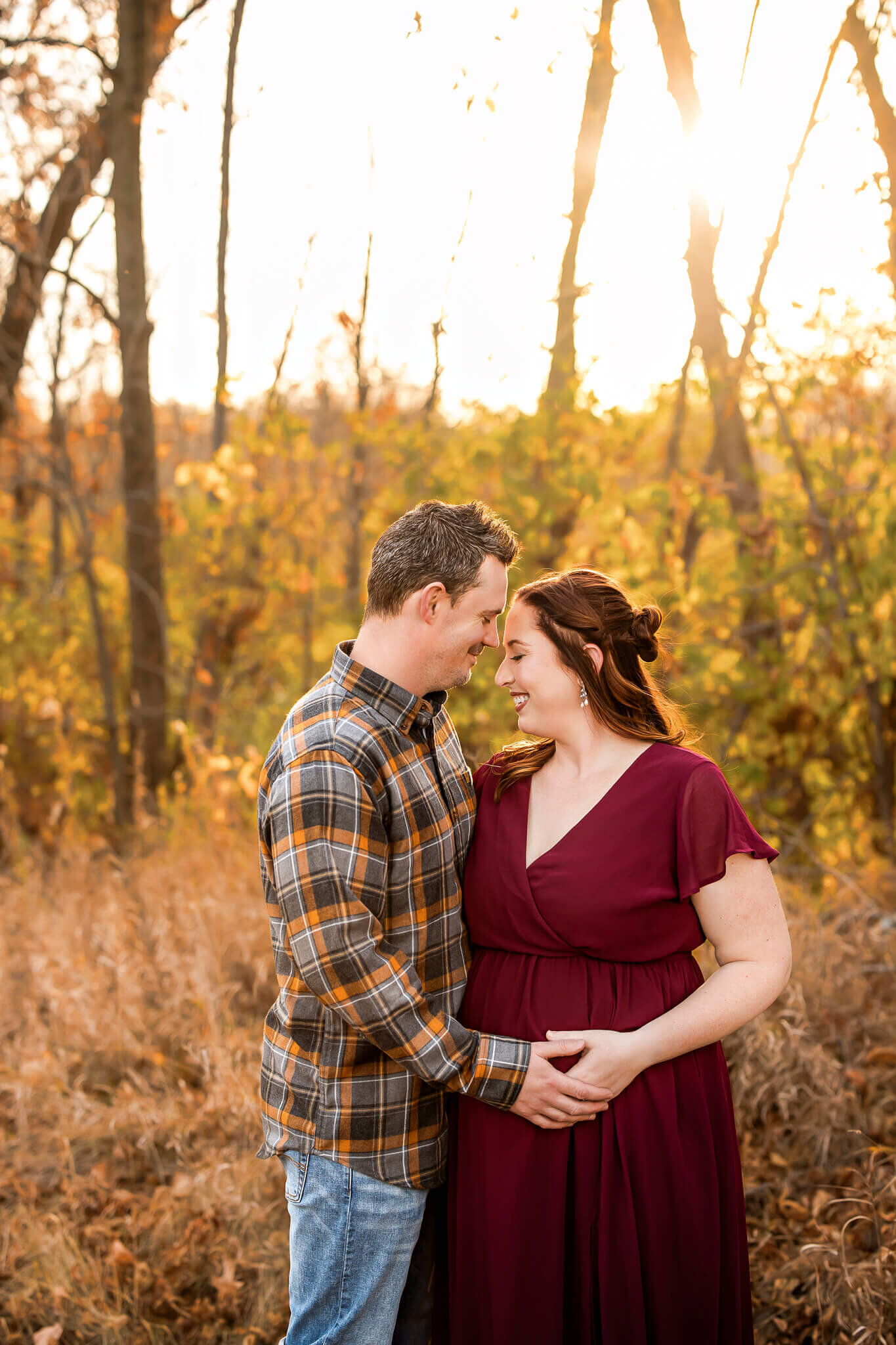 A couple stands in a sunlit autumn field, smiling at each other. The woman in a burgundy dress holds her baby bump while the man gently embraces her—perfect for maternity portraits by a talented Bloomington maternity photographer.