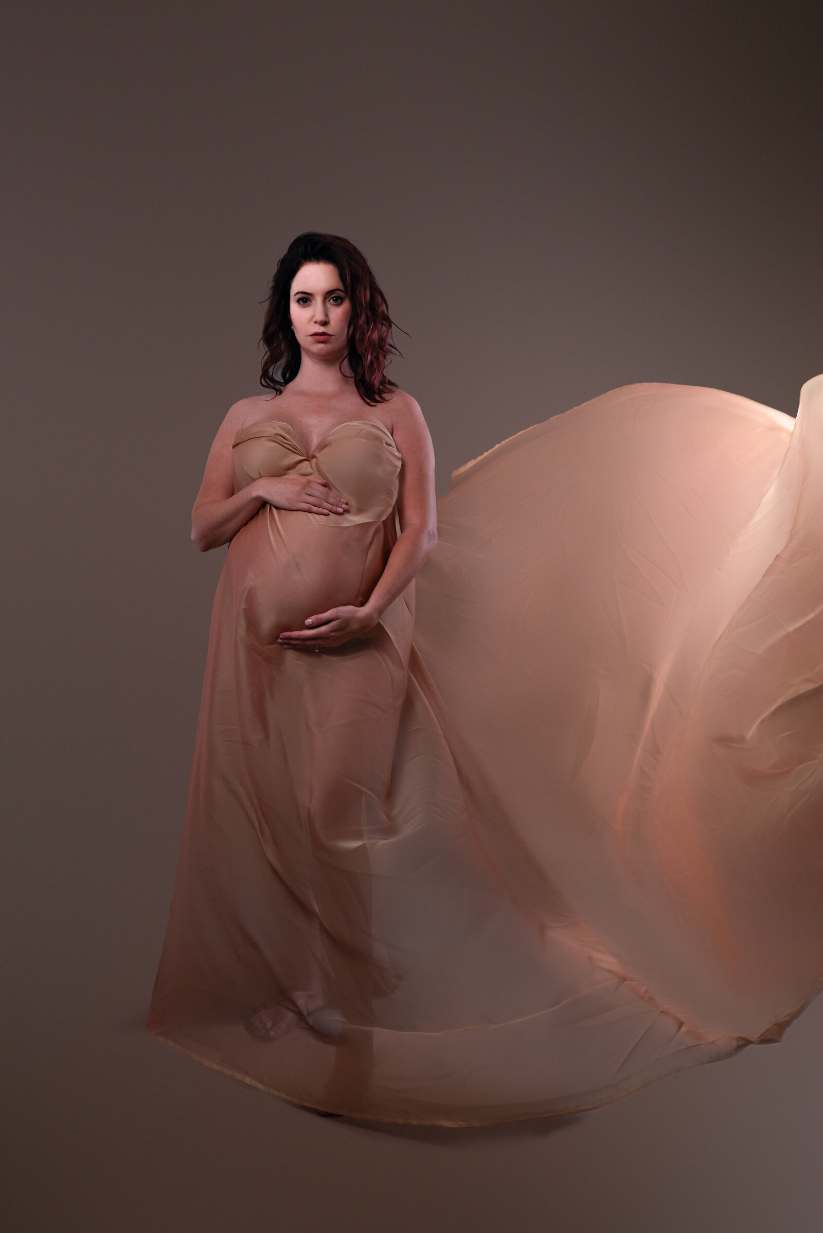 A pregnant woman wrapped in  a flowing gold fabric for her studio maternity pictures in Huntsville Alabama