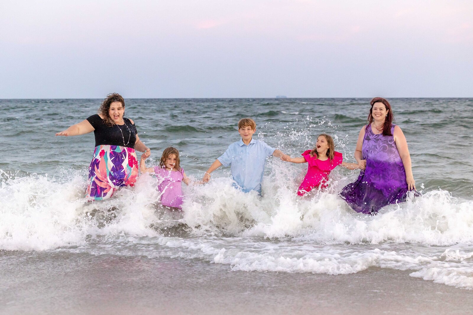 A family joyfully plays in the ocean waves at sunset, wearing colorful outfits. The lively scene captures the essence of fun and connection, ideal for artistic family photography in Wilmington, NC, showcasing playful beachside interactions.