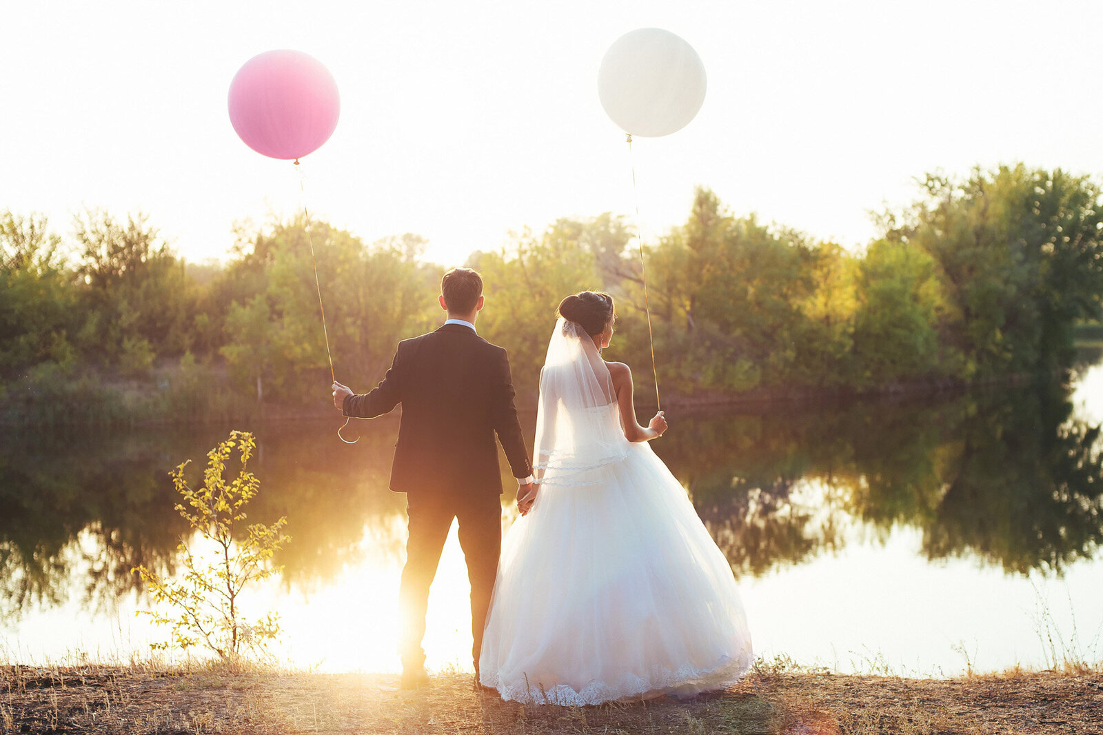 Bride holding white balloon and groom holding pink balloon while holding each other's hand facing the lake