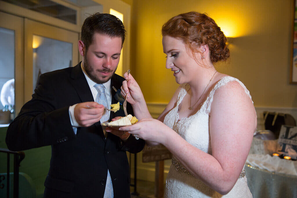A bride and groom, dressed in wedding attire, smile and feed each other cake indoors, celebrating their wedding reception.