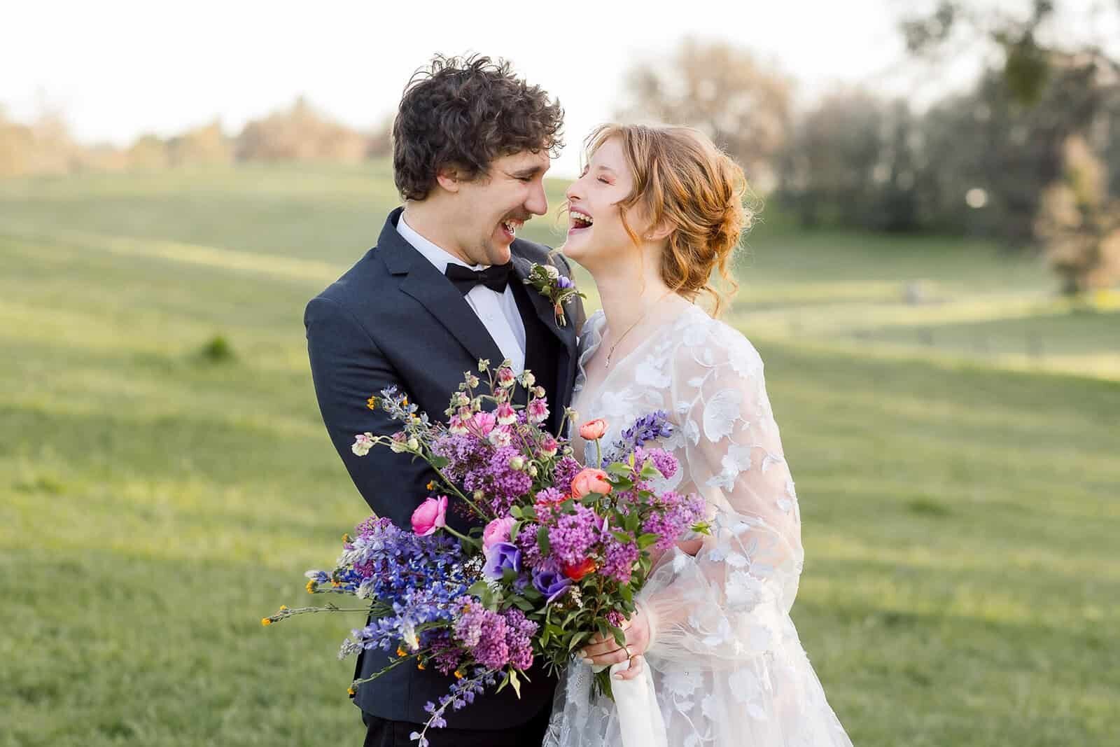 A bride and groom laugh together while standing in a lush green meadow on their wildflower themed wedding day. 
