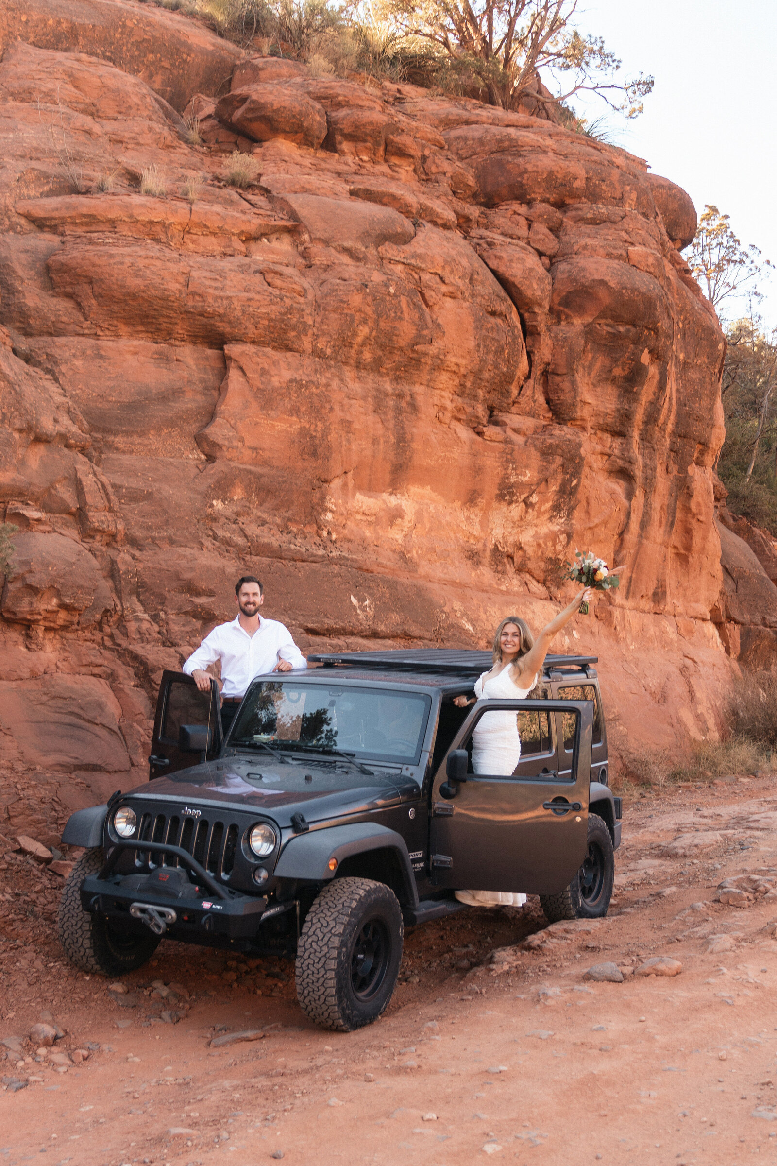 Bride and groom celebrating their adventurous elopement on the rugged trail to Merry Go Round Rock in Sedona, Arizona, standing in and around a black Jeep with red rock cliffs in the background.