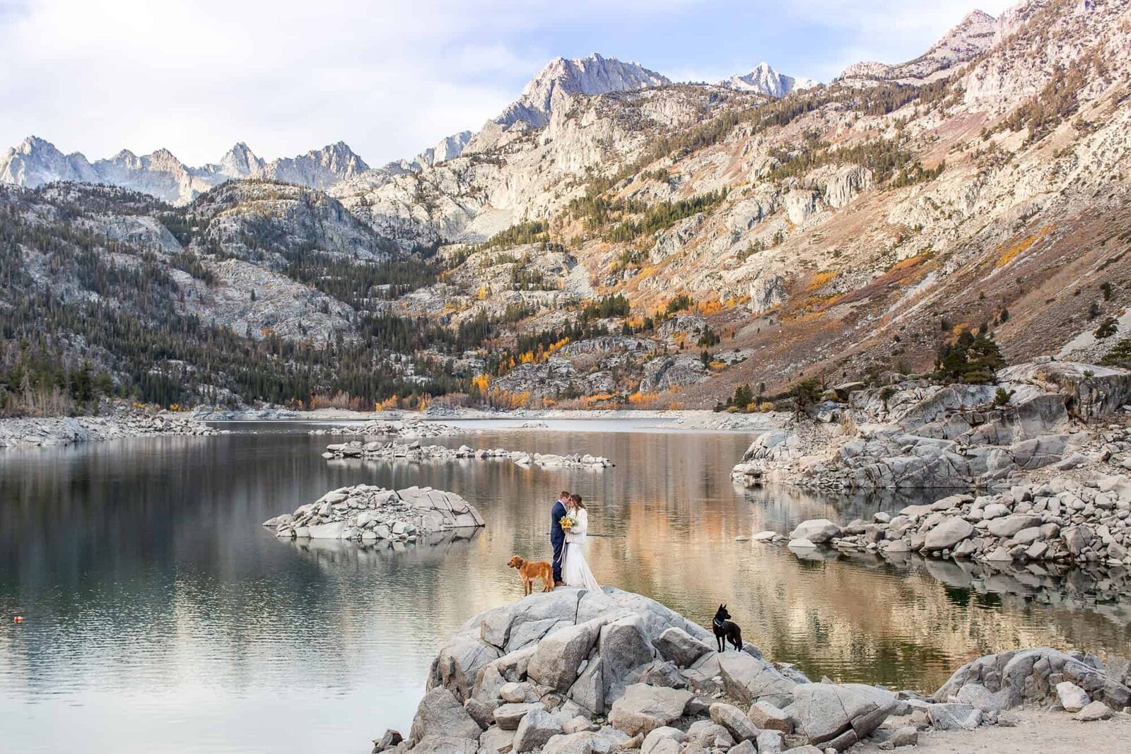 A bride and groom enjoy their wedding day with their two dogs, in front of a pristine alpine lake in the Eastern Sierras at sunrise.