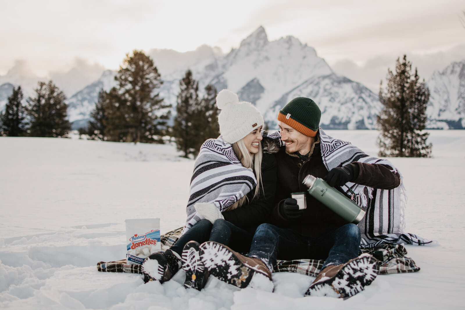 photographers jackson hole photographs darling engagement session in the mountains with man and woman cuddled up under a blanket together with hot chocolate as they laugh together surrounded by snow with woods behind them