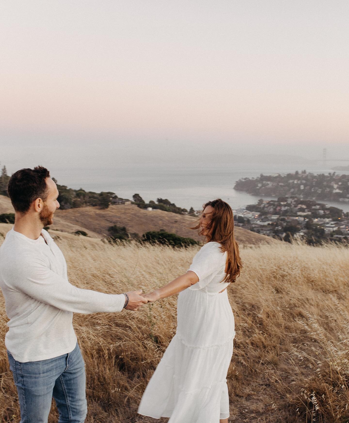 A couple holds hands in a golden field overlooking a scenic coastline at sunset, creating a warm, peaceful ambiance.