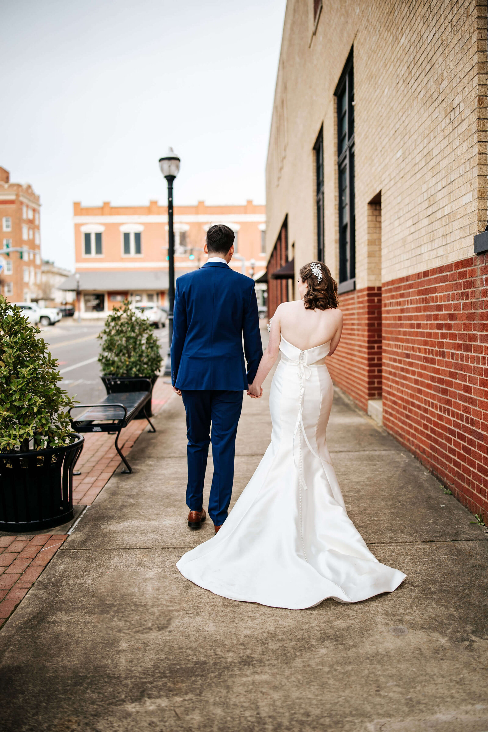 A bride in a white gown and a groom in a blue suit walk hand-in-hand down a city sidewalk, away from the camera, beside a brick building on a bright day.