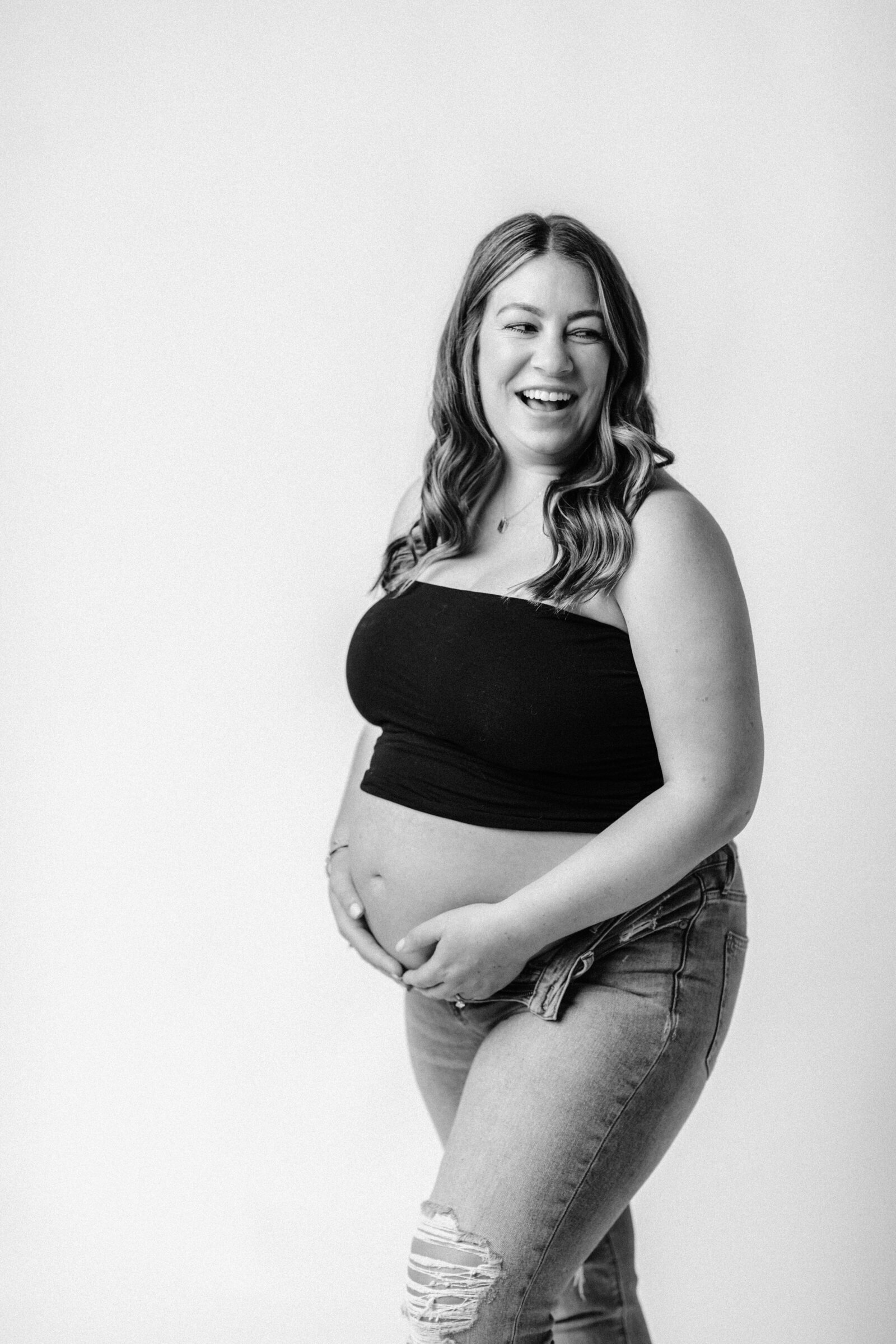 pregnant mother in black tube top and jeans holding baby bump laughing 