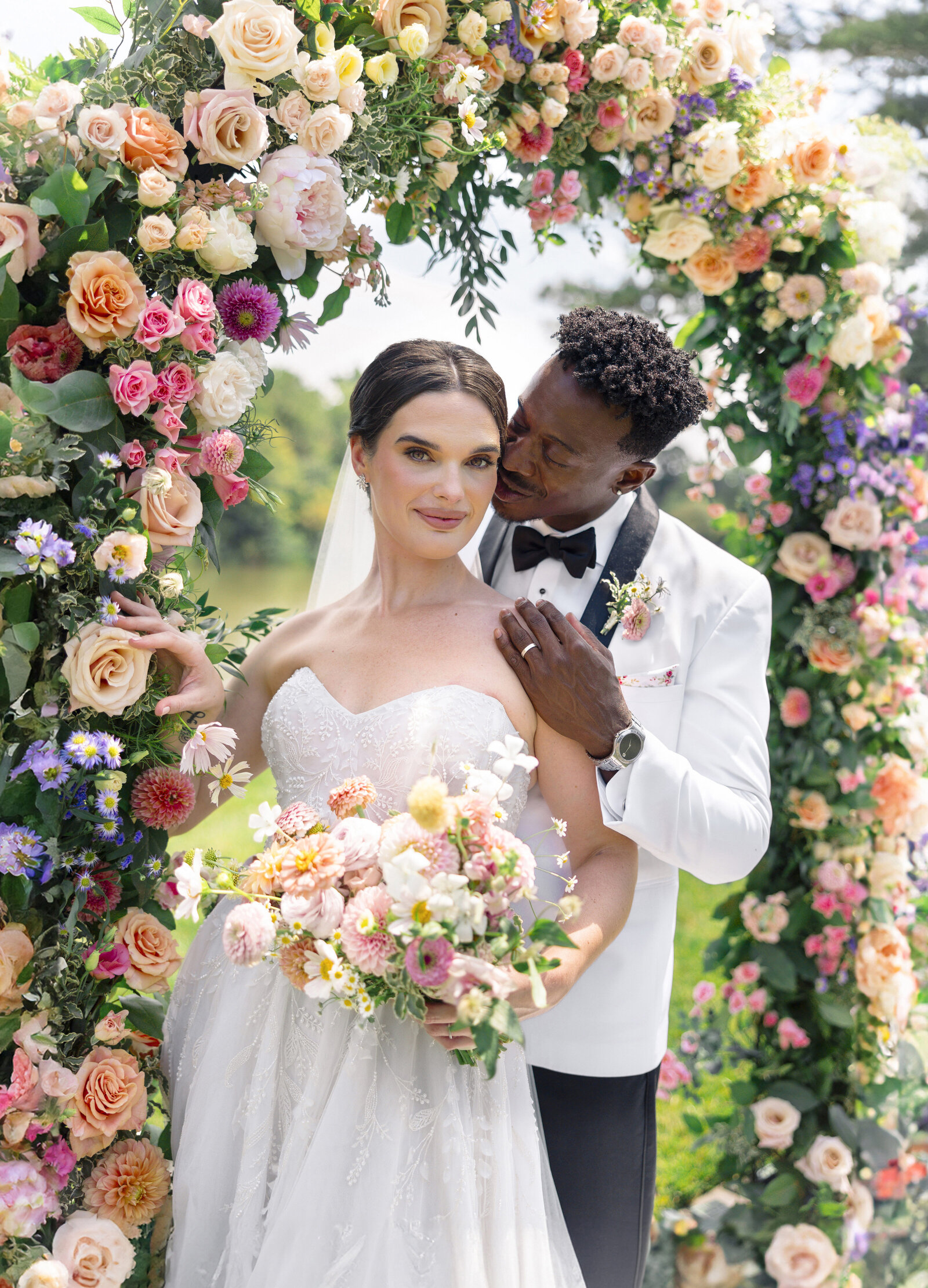 A bride and groom pose under a lush floral arch. The bride wears a strapless white gown, holding a bouquet of roses and daisies. The groom, in a white suit and black bow tie, stands behind her gently. The arch is adorned with vibrant pink, peach, and white flowers.