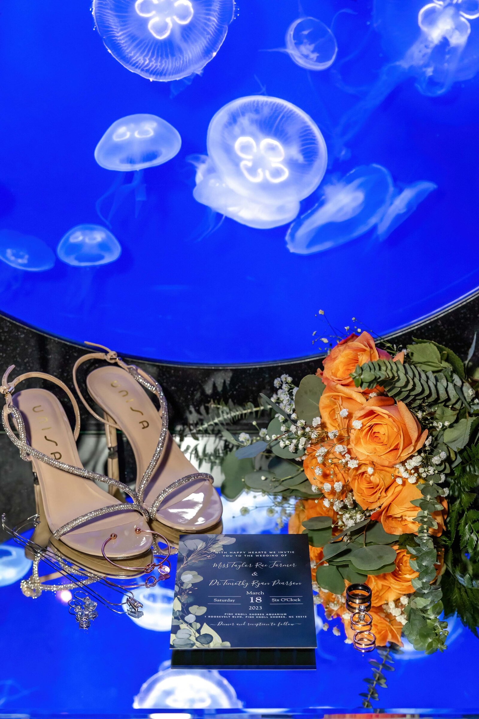 A bridal flat lay featuring sparkling heels, orange roses, wedding invitations, and a glowing jellyfish exhibit backdrop, captured during a Pine Knoll Shores, NC wedding, blending elegance with marine-inspired creativity.