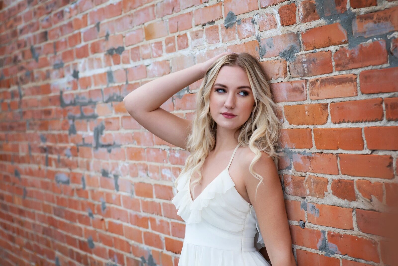 A blonde standing against a brick wall in Akeley, Minnesota wearing a white dress looking off into the distance.