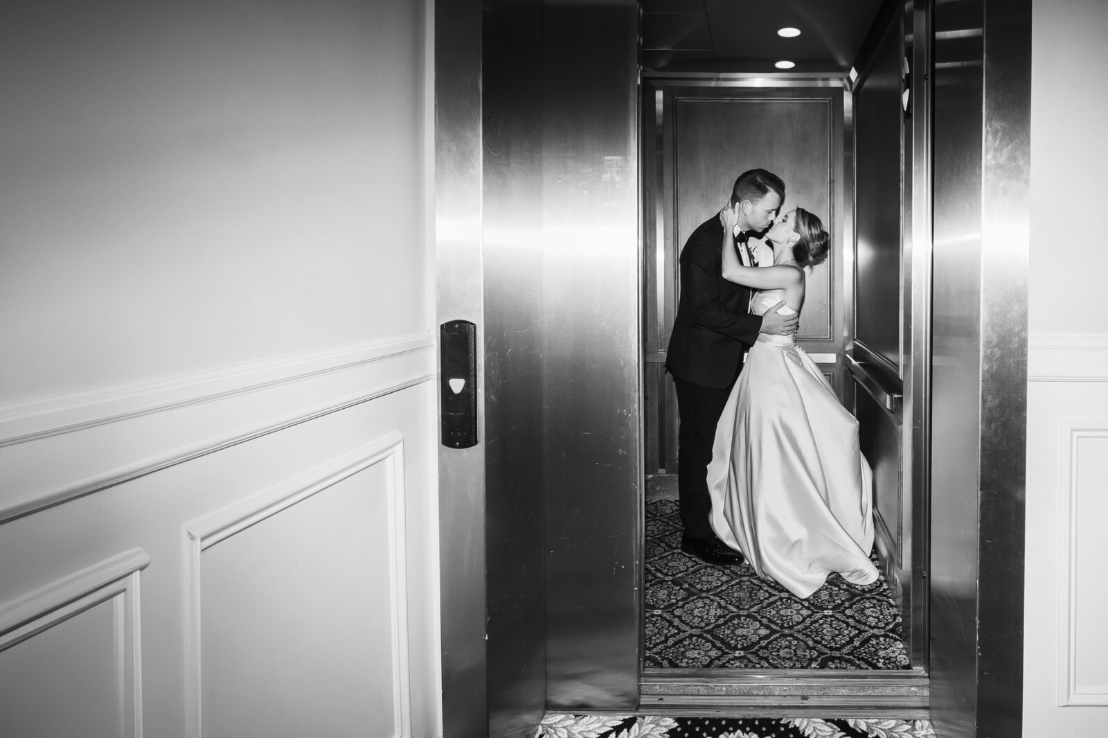 A couple in formal attire kisses inside an elevator with open doors. The setting is elegant, with a patterned floor and paneled walls. The lighting is soft and intimate.