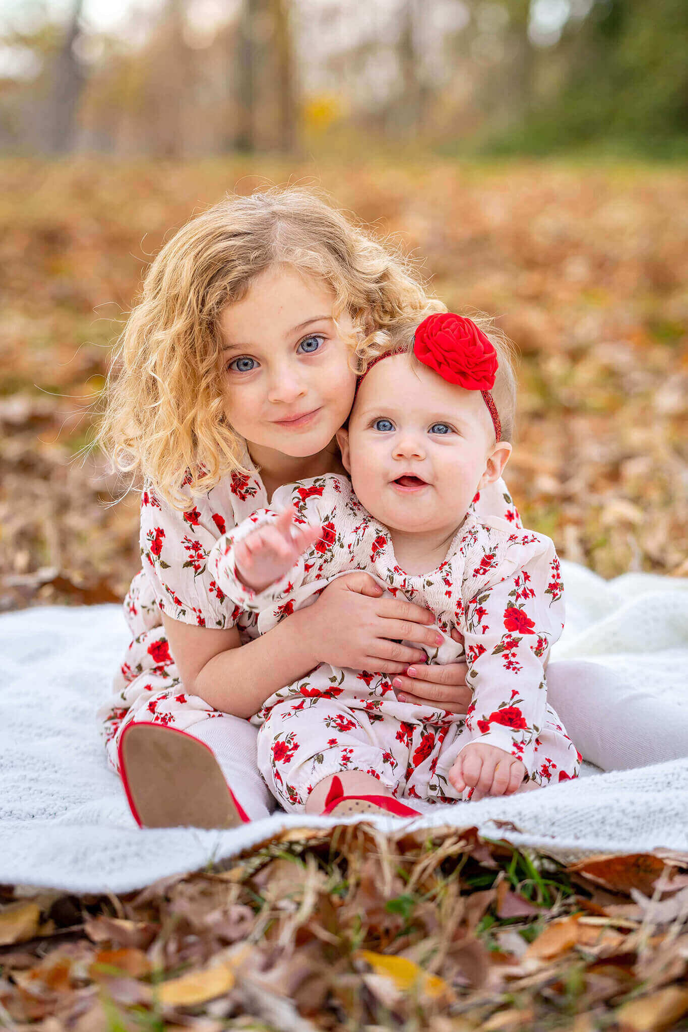Two girls sitting on a while blanket surrounded by fall leaves.