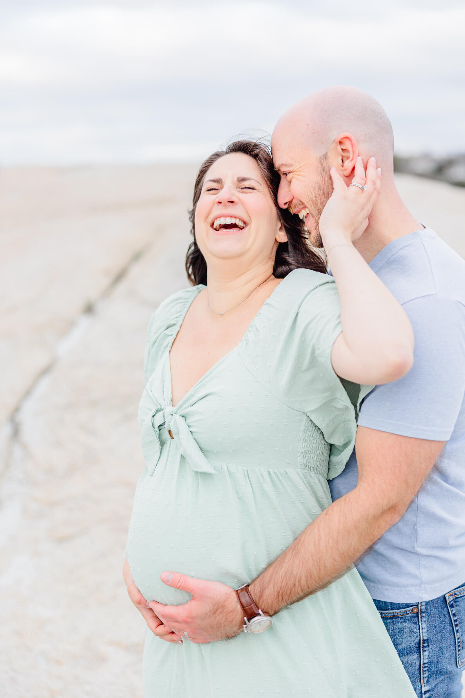 Pregnant woman in a flowy mint dress laughs heartily with her husband as he embraces her baby bump from behind at a Northshore Boston maternity photo session