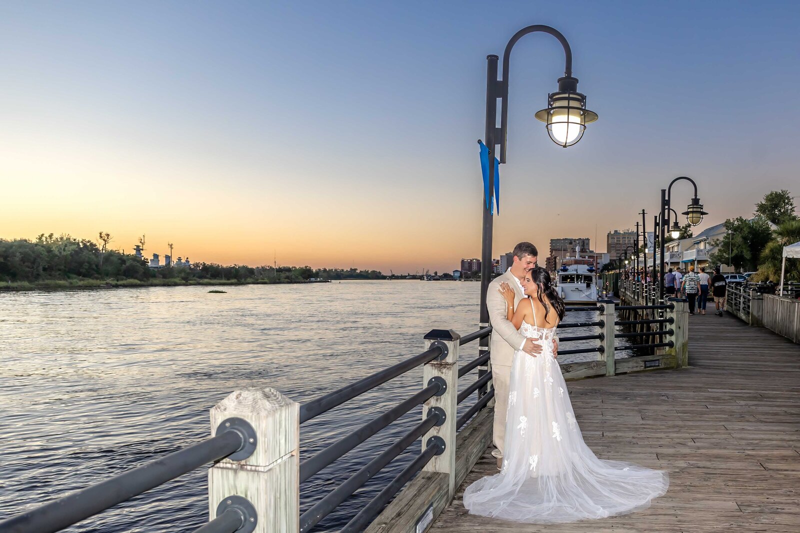 The bride and groom embrace on the Riverwalk at sunset, with the Cape Fear River and city lights in the background, captured during a Wilmington, NC wedding, blending romance and coastal charm.