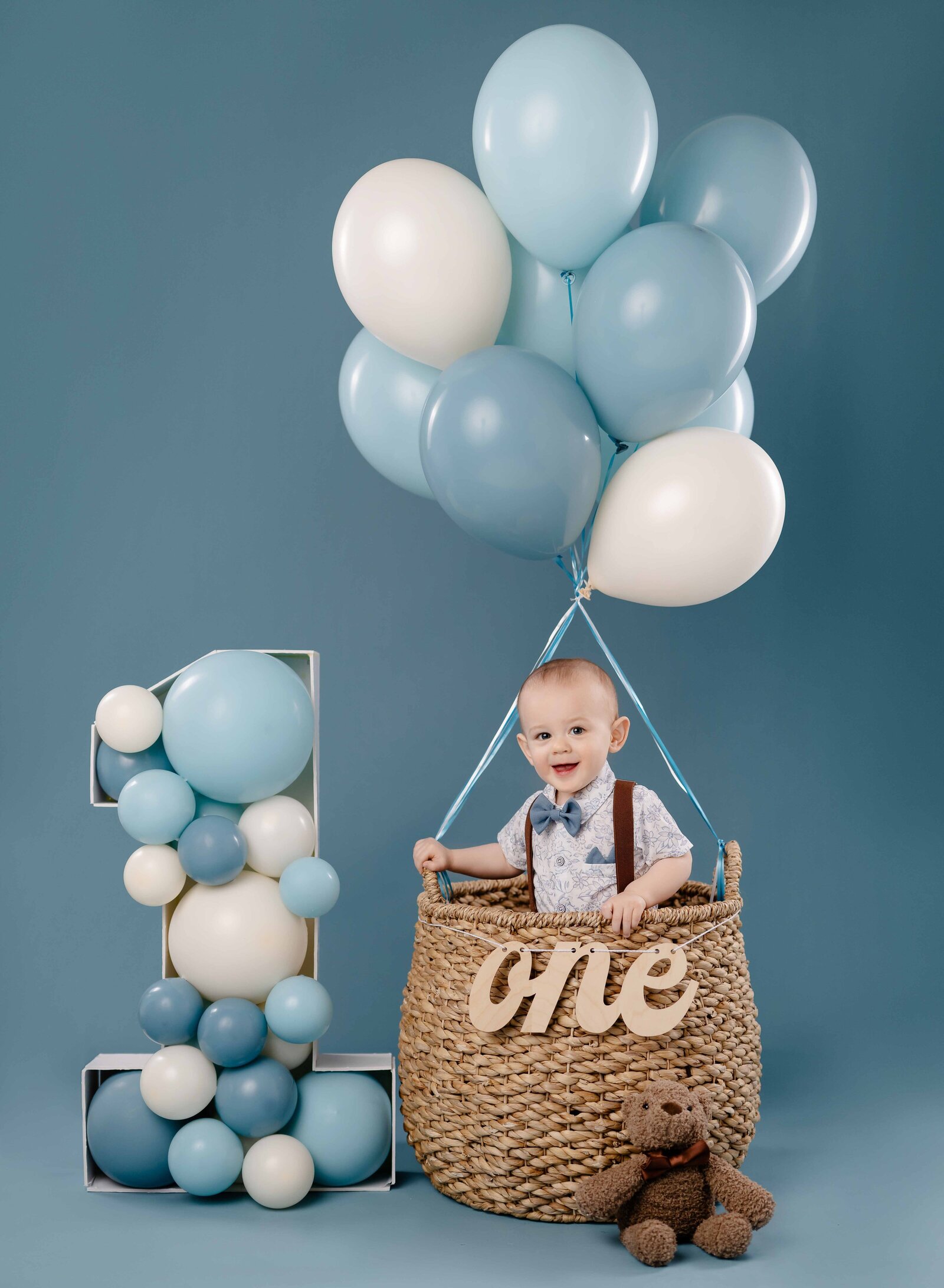 Baby standing in balloon basket smiling with balloon filled #1 marquee next to him. 