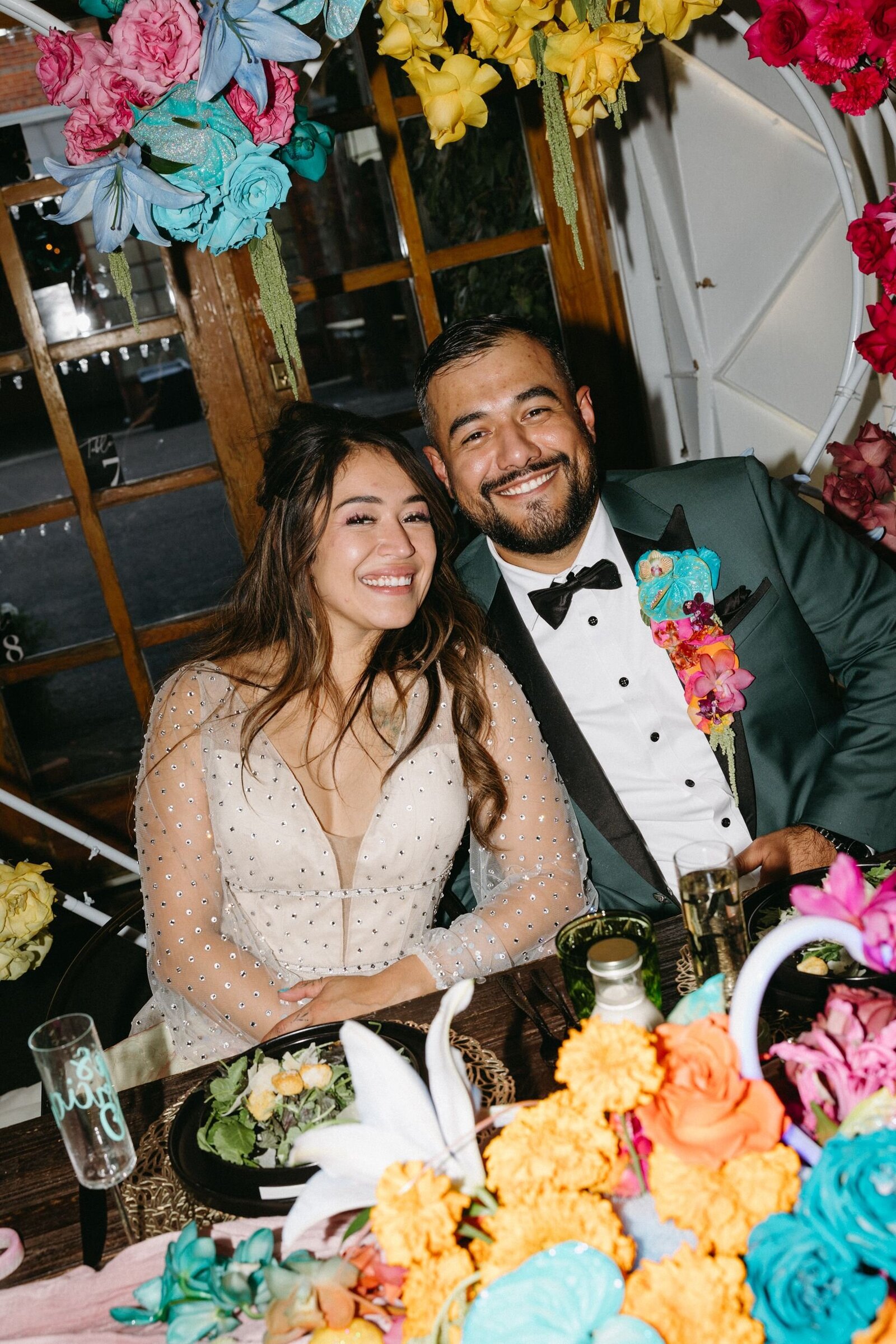 A bride and groom sit at their sweetheart table surrounded by colorful florals