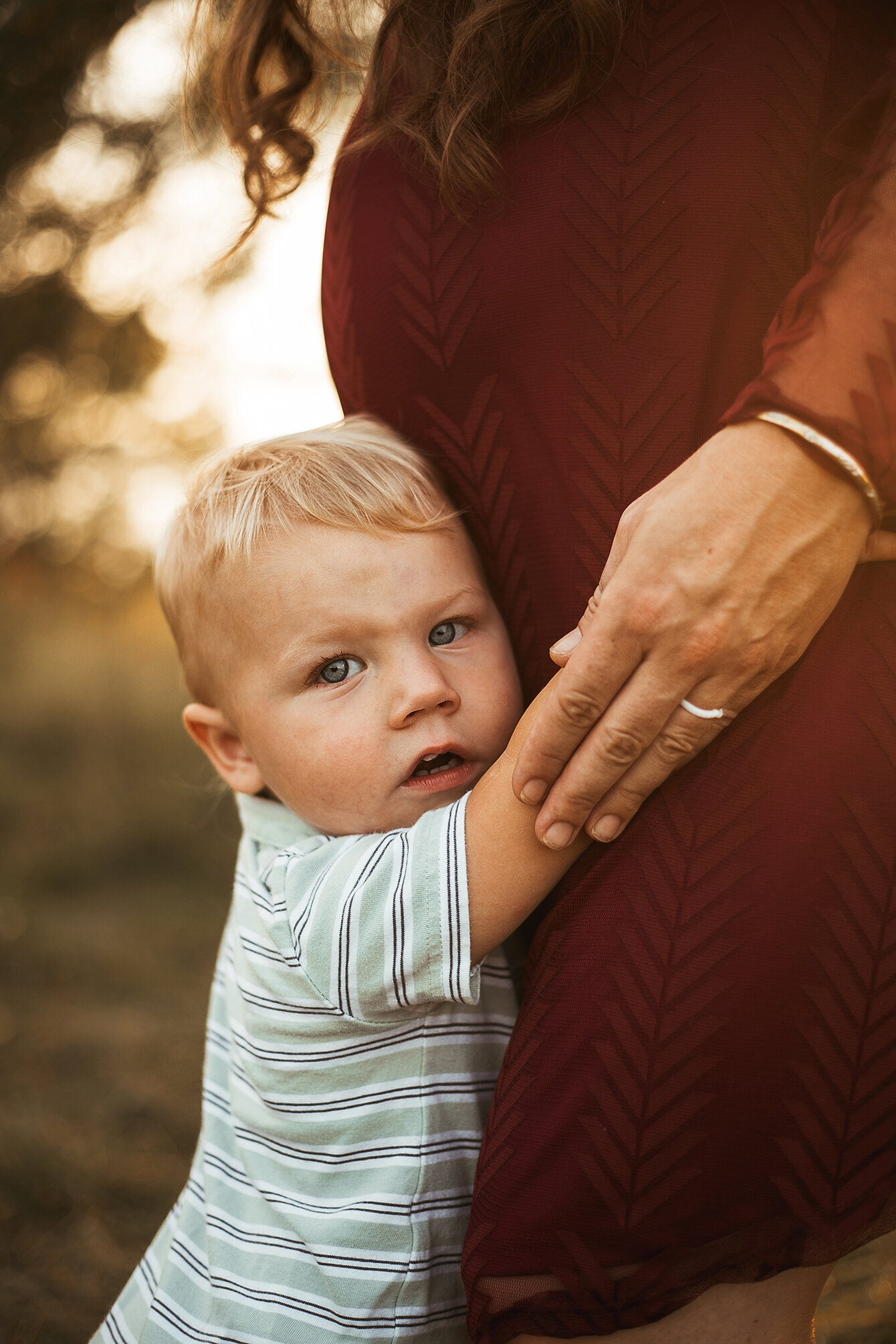 Toddler hugging and leaning into mommy’s pregnancy belly. Golden light in background. Child looking at the camera.  Kids Photography. Maternity Photography.