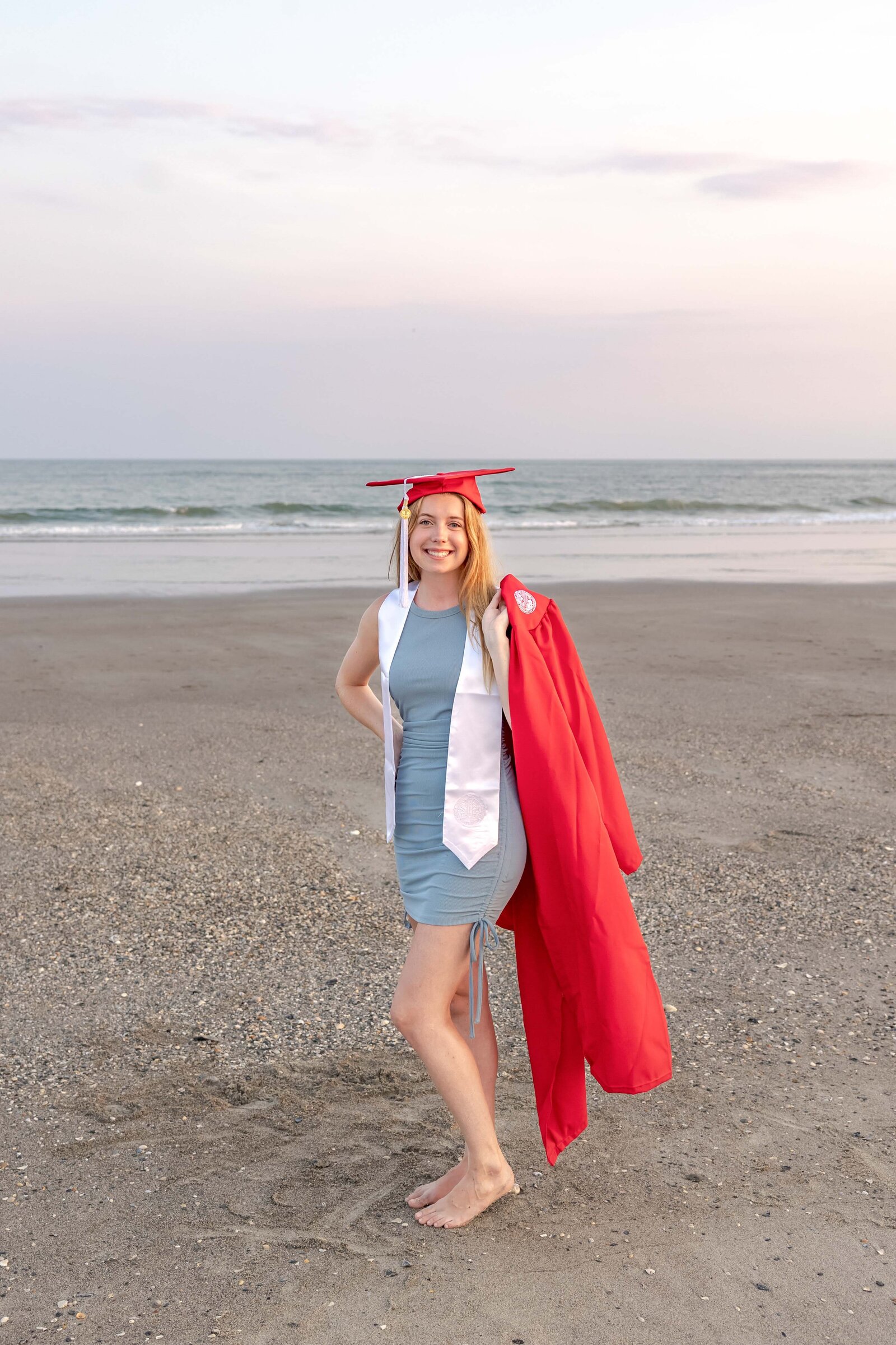 A college senior in a red cap and gown standing barefoot on the beach with the ocean in the background in Oak Island, North Carolina. This joyful image captures the celebration of graduation, perfect for capturing memorable senior moments by the sea.