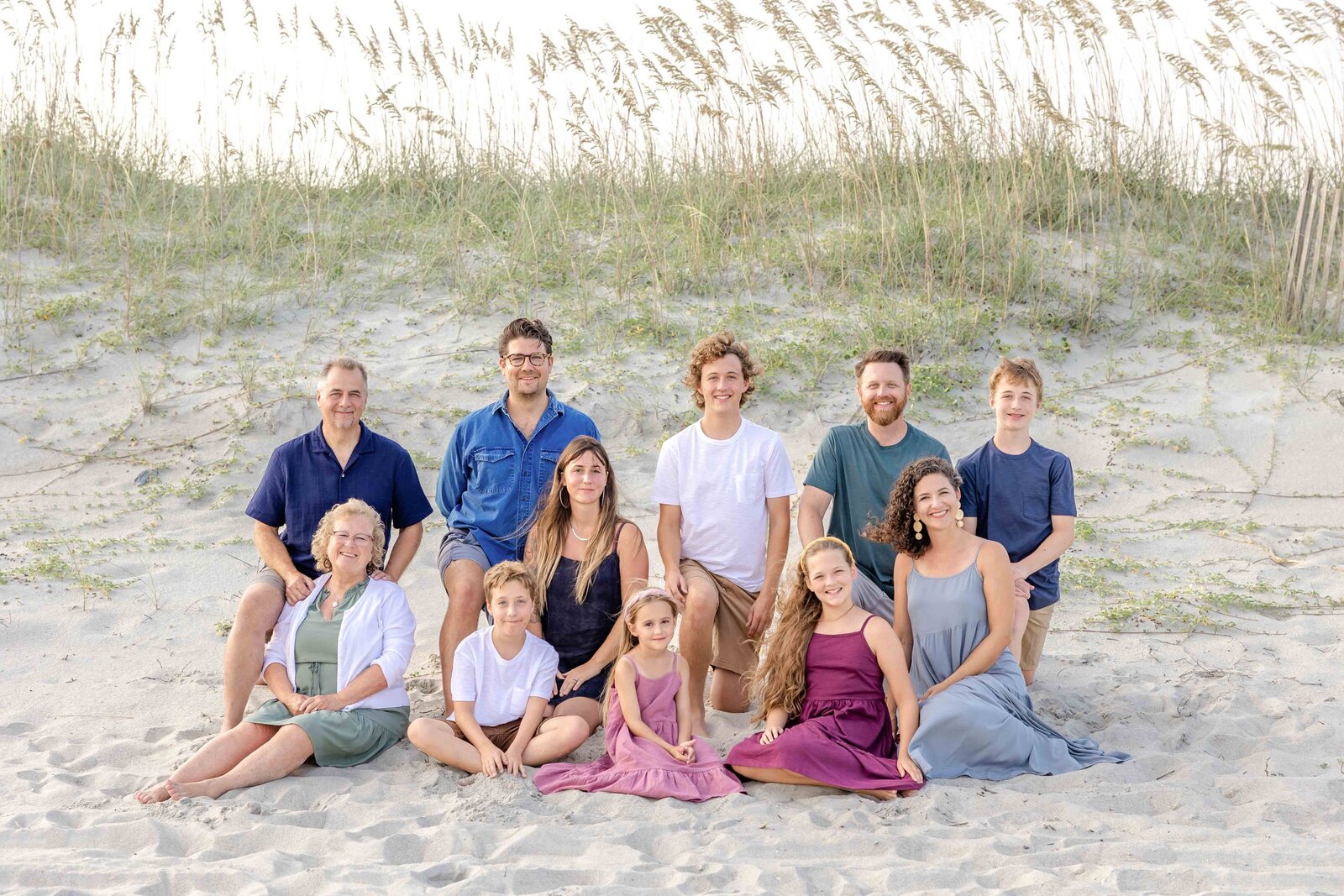 An extended family sits on the sandy dunes at Ocean Isle Beach, surrounded by sea oats. Dressed in a mix of blues and purples, they capture the essence of a relaxed family gathering. Perfect for destination family photographer NC, highlighting scenic coastal settings.