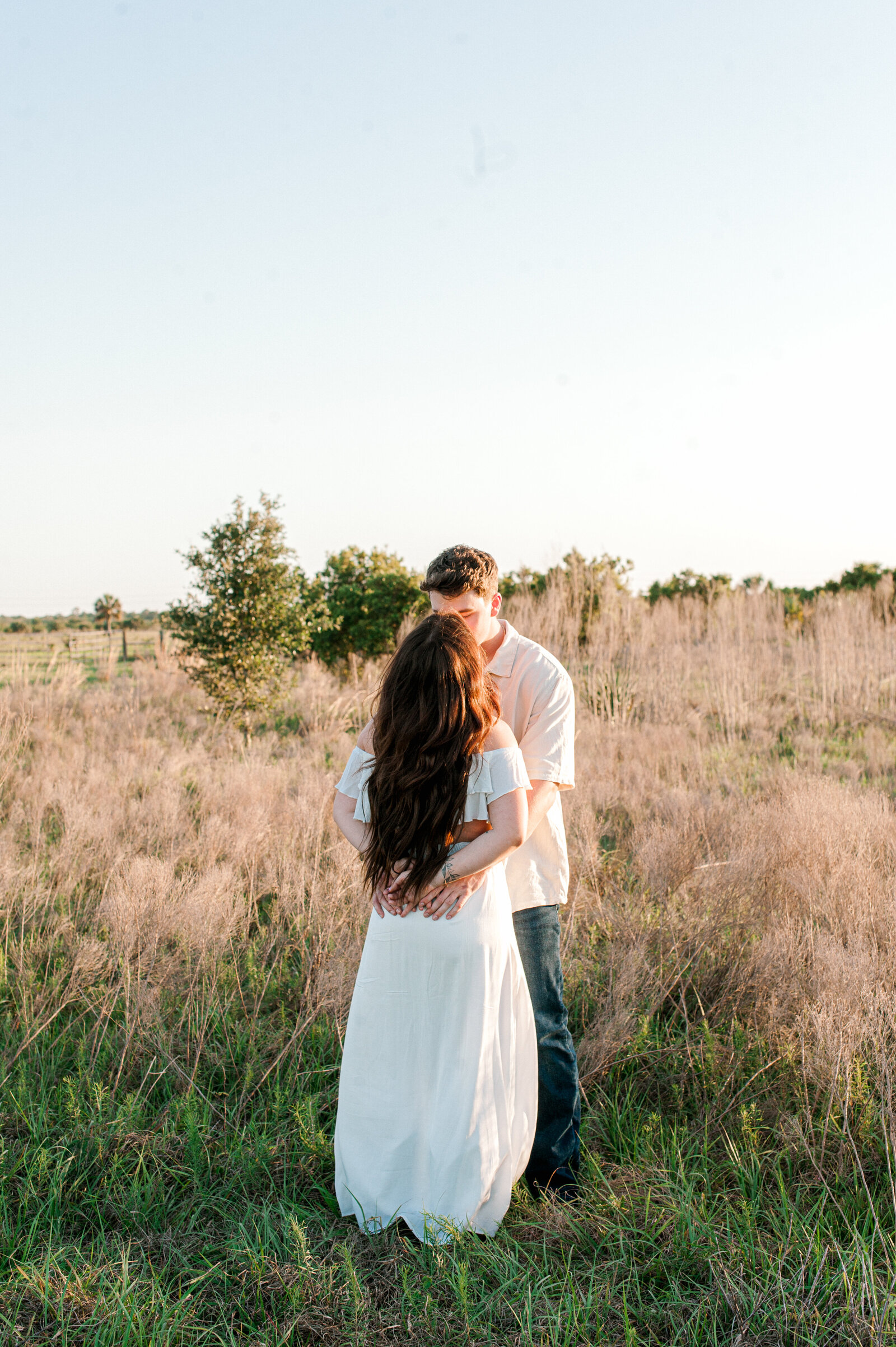 Couple stands in a tall grass field kissing at golden hour