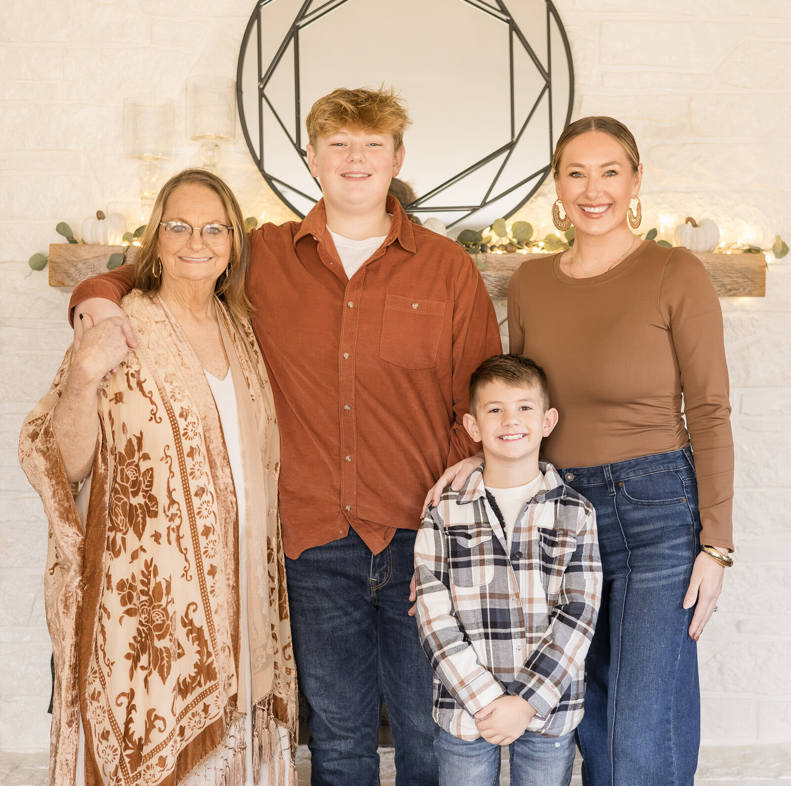 A grandma, mom, and two sons standing in front of a fireplace smiling at the camera.  Grandma has blonde brown hair, is wearing a tan and red smock with a white shirt under it.  The older son has brown hair wearing a dark red button up with a white shirt under it and blue jeans.  Young son has black hair, wearing a flannel button up with a white shirt under it, and blue jeans.  Mom is wearing a long sleeve tan shirt and blue jeans with brown hair.