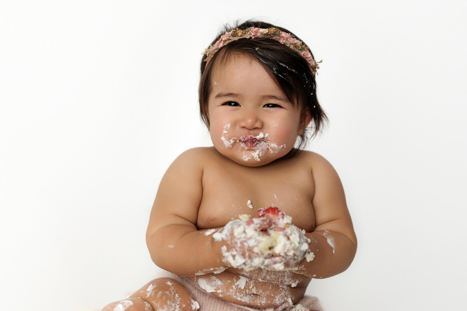 A happy baby with dark hair, wearing a floral headband and a pink diaper cover, sits on a white backdrop. The baby is joyfully smashing a cake, with frosting smeared all over their face, hands, and arms, and a big smile showing their delight. The scene captures the fun and messy moments of a cake smash session.