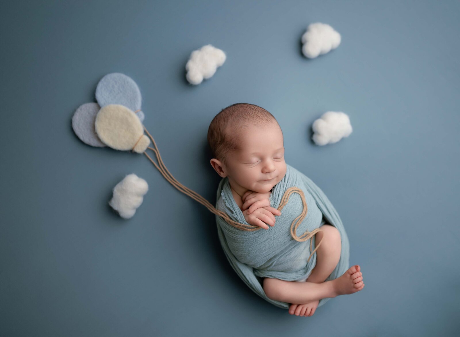 Baby boy on blue backdrop, holding felted balloons and surrounded by clouds. Photo taken by Jenni Nikki at Newborn photo studio in Atlanta, GA.