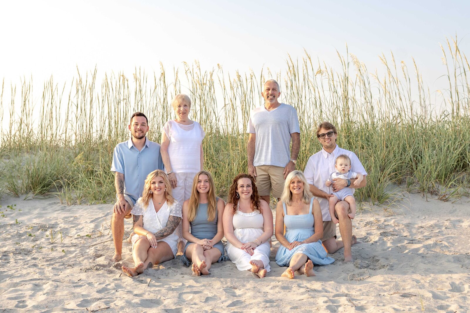 A multi-generational family sits together on the sandy shore of Oak Island, dressed in soft pastel colors. With sea oats swaying behind them, they capture the warmth of a family gathering. Ideal for those seeking the best family photographer in North Carolina for timeless portraits.