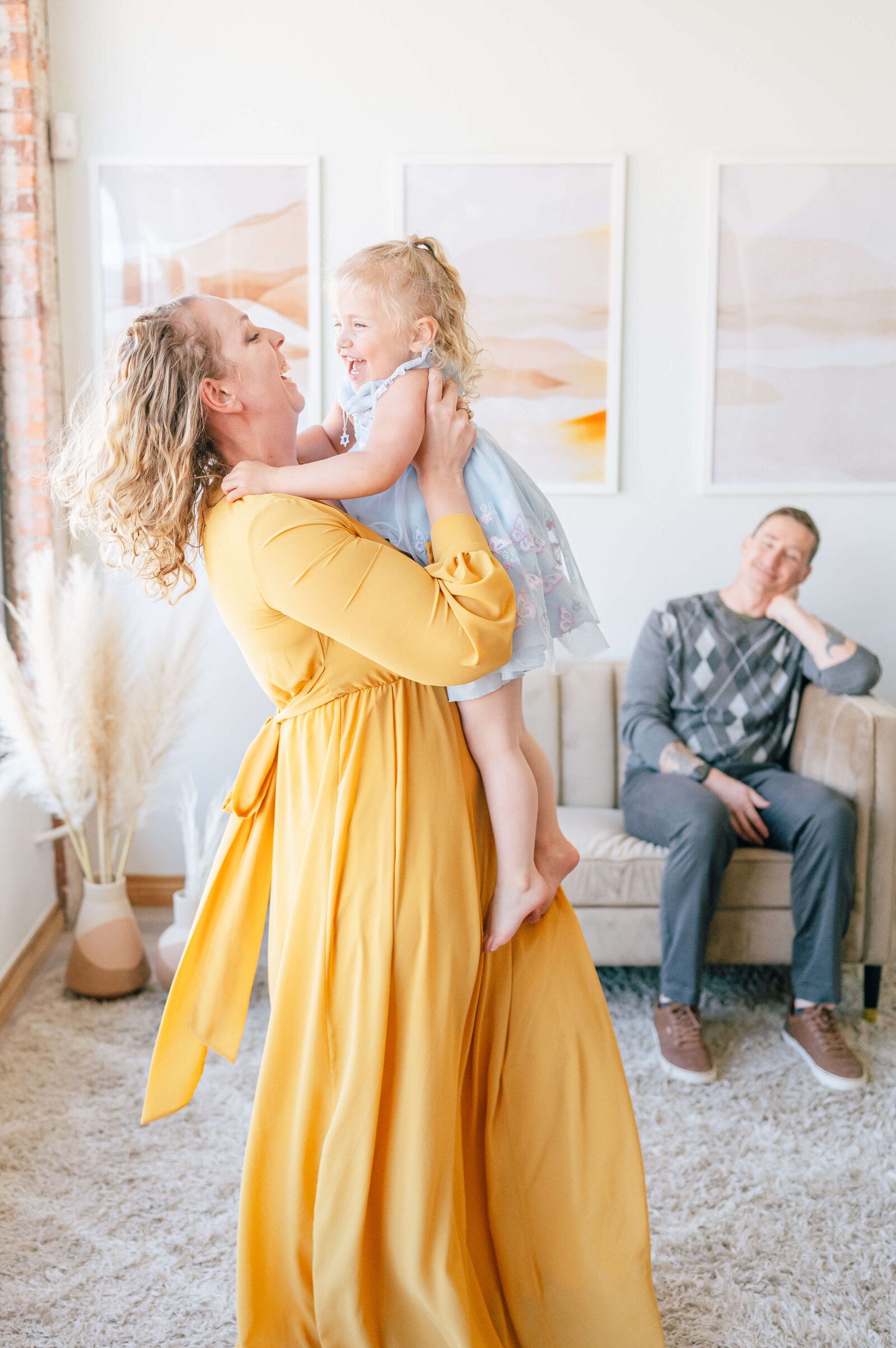 A mother with curly blonde hair wears a flowing yellow dress and lifts her young daughter in the air while exhibiting a big and happy smile