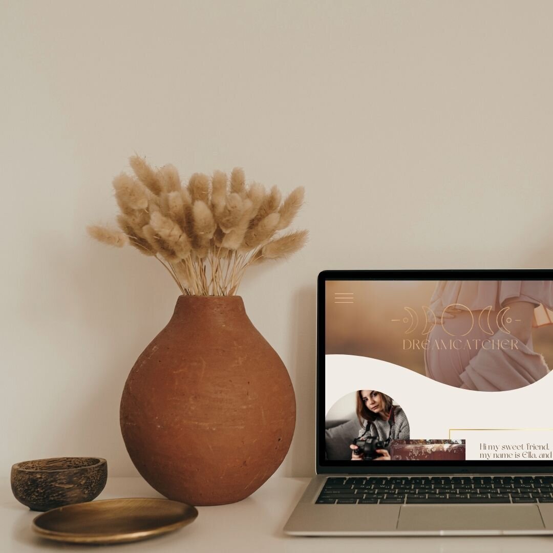 A minimalist workspace with a laptop, ceramic vase with dried pampas grass, and a wooden bowl on a neutral background.