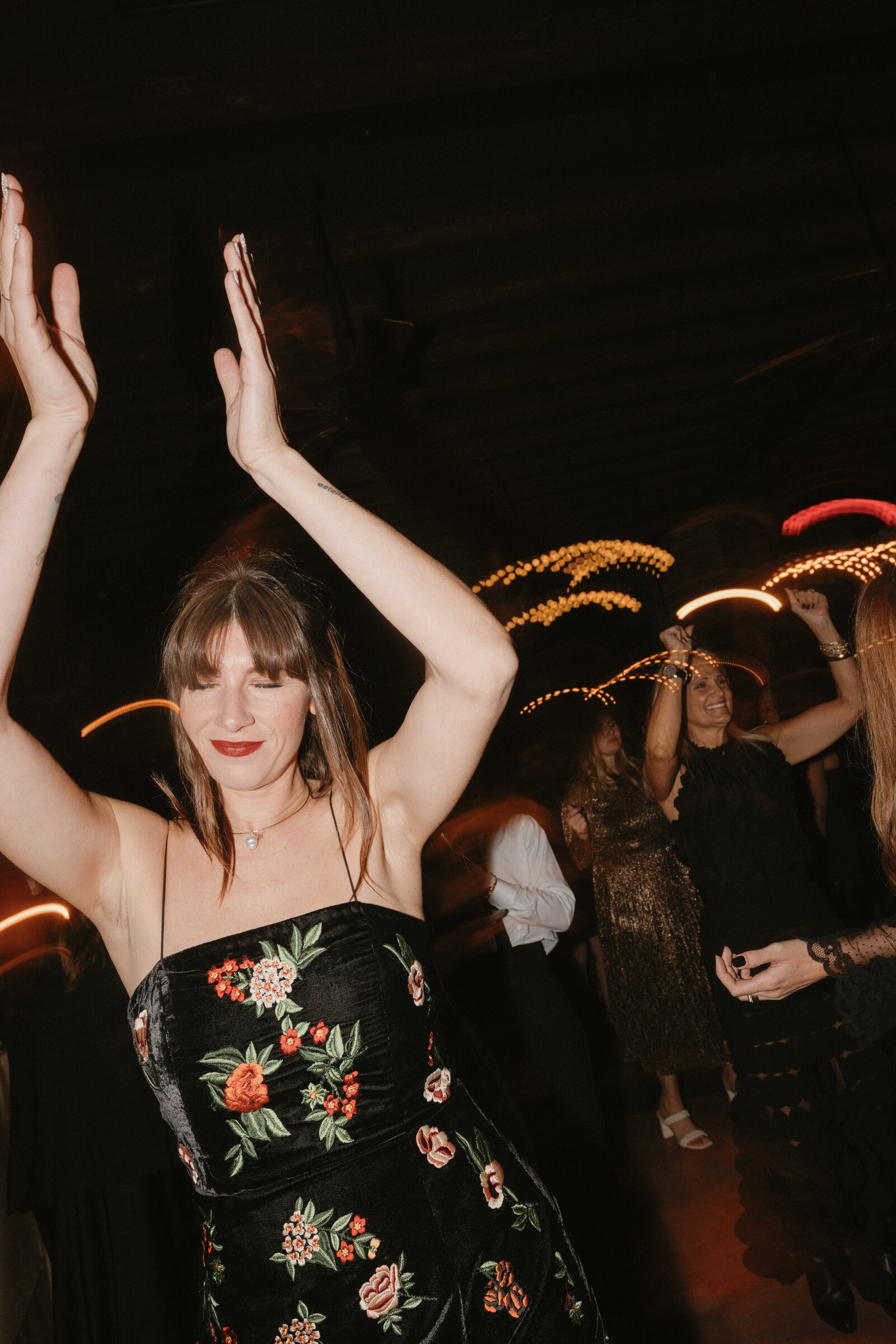 Direct flash photo of female wedding guest with bangs in black velvet floral spaghetti strap dress clapping hands above head on dance floor 