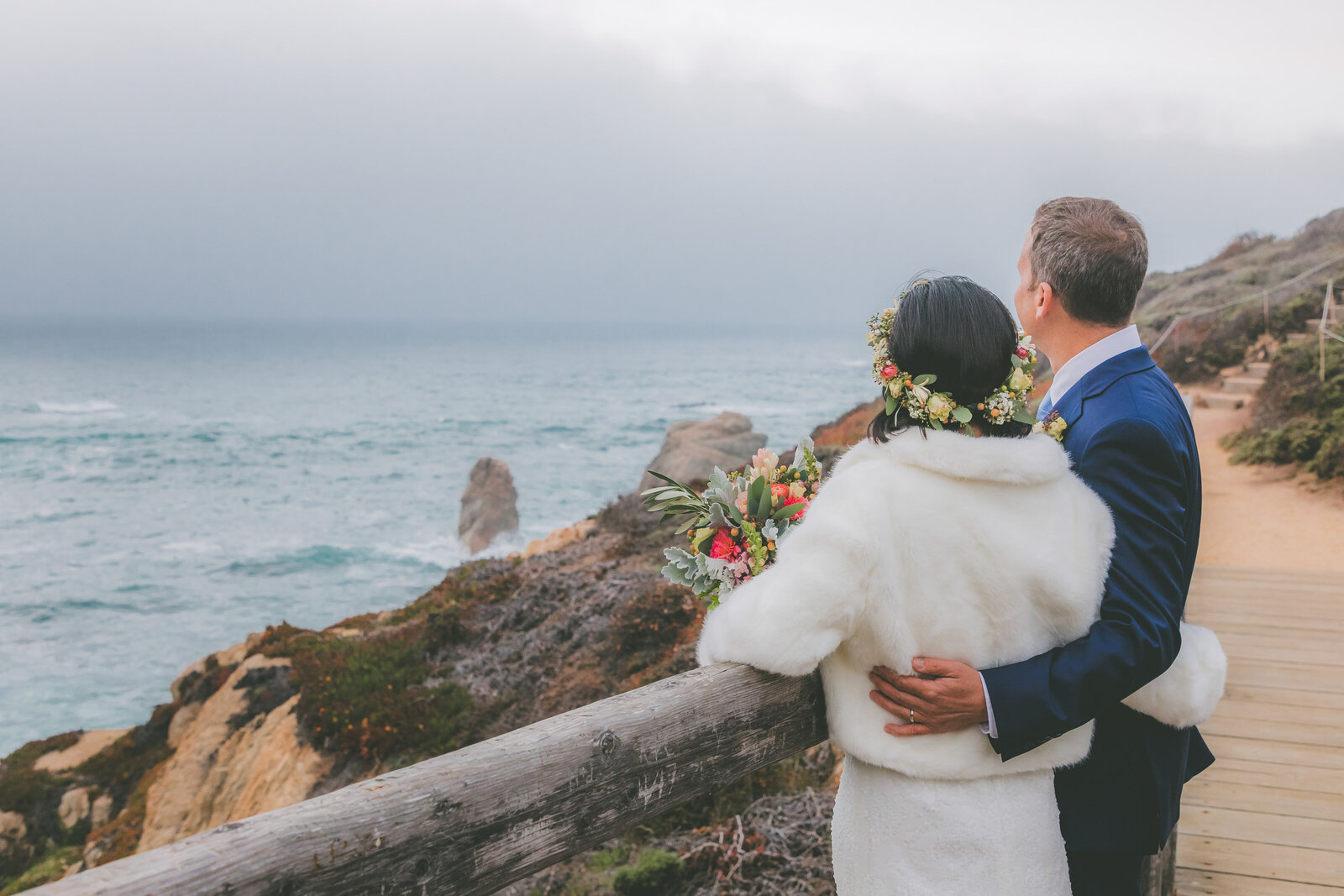 You can see the backs of a bride and groom as they hug each other and stare into the ocean view in Big Sur.